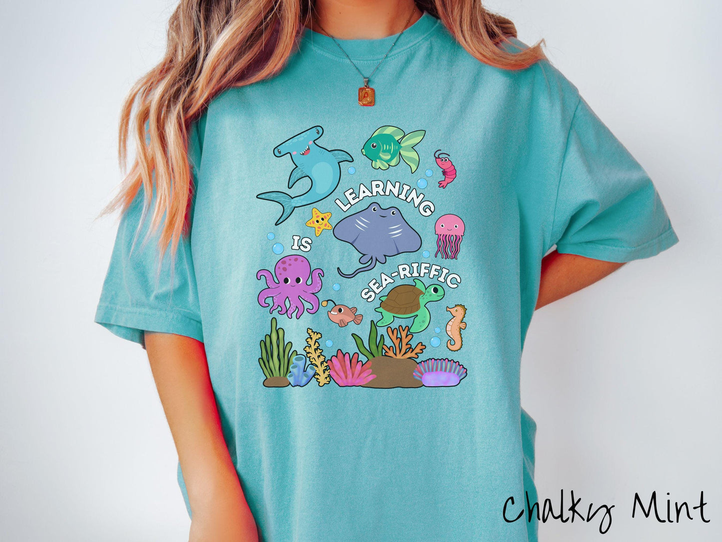 A woman wearing a cute, vintage chalky mint colored Comfort Colors T-shirt with the text Learning is Sea-Rific. There is a shark, green fish, octopus, stingray, jellyfish, turtle, and seahorse swimming together above a colorful coral reef.