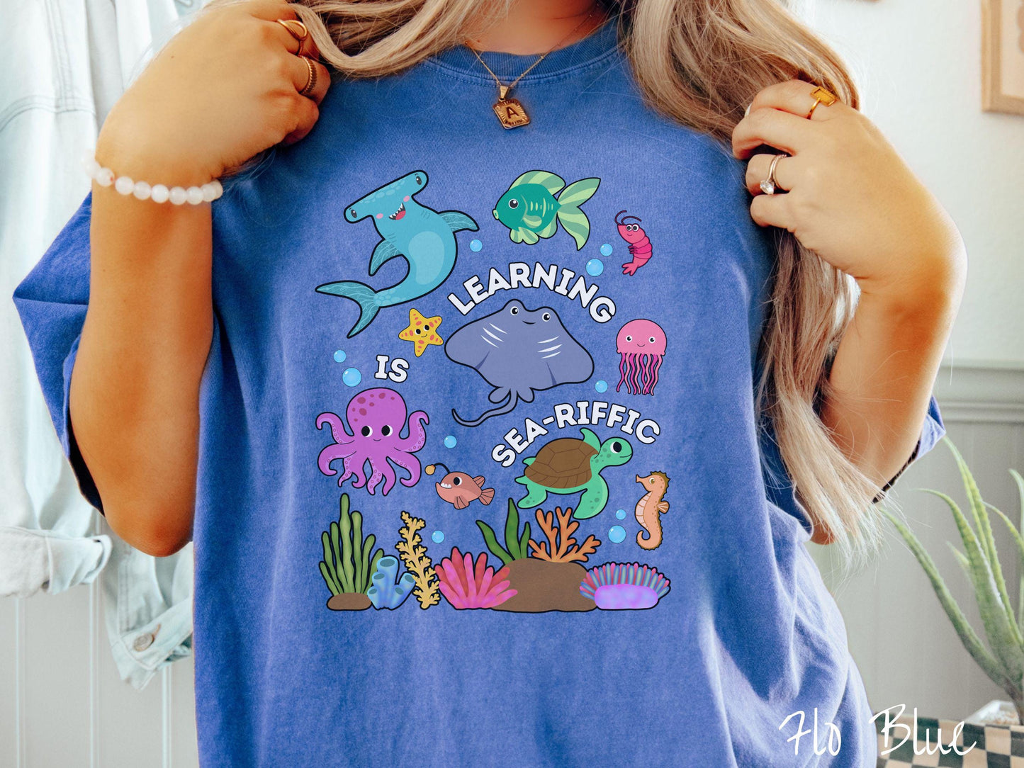 A woman wearing a cute, vintage flo blue colored Comfort Colors T-shirt with the text Learning is Sea-Rific. There is a shark, green fish, octopus, stingray, jellyfish, turtle, and seahorse swimming together above a colorful coral reef.