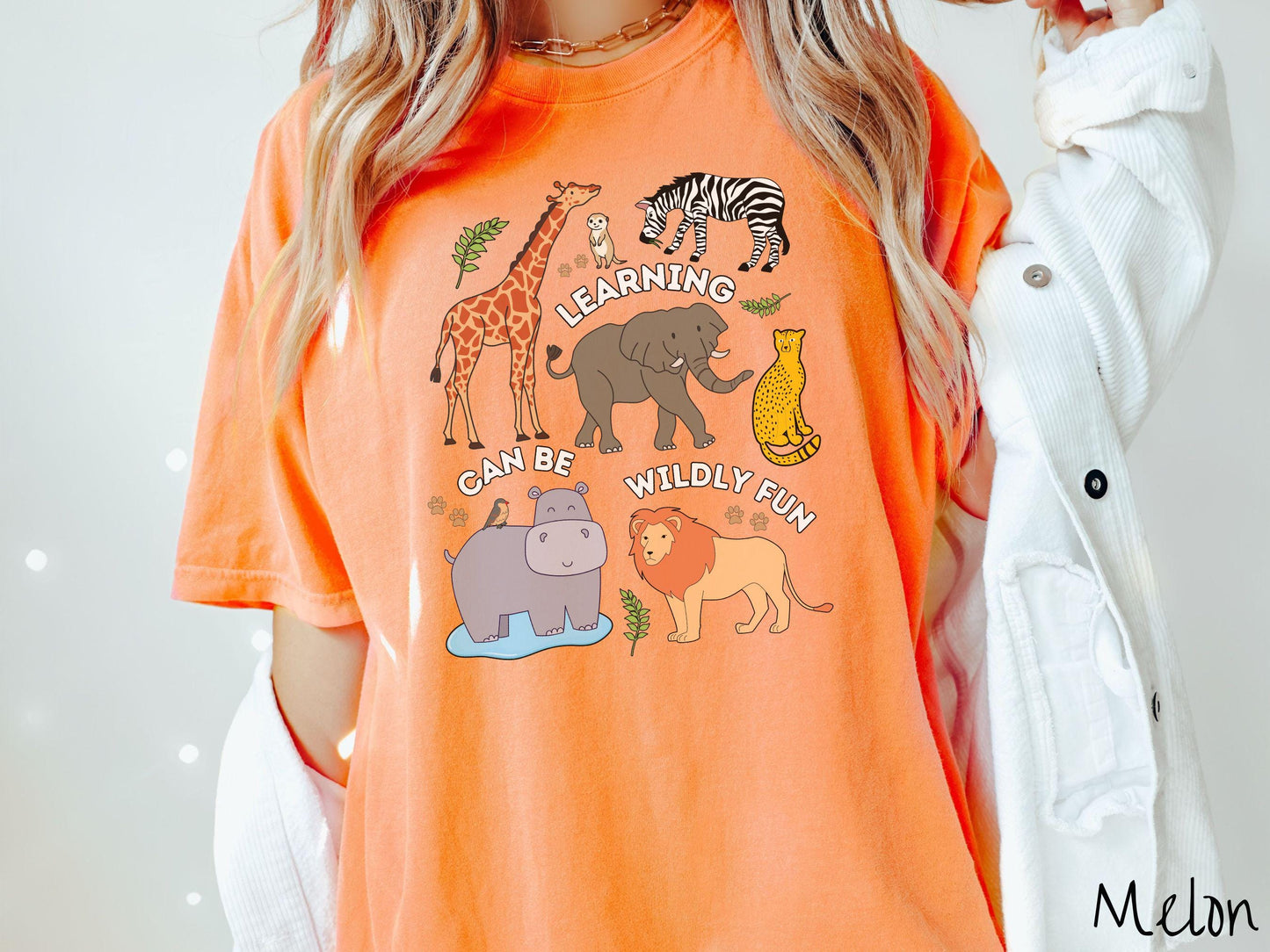 A woman wearing a cute, vintage melon colored Comfort Colors T-shirt with the text Learning Can Be Wildly Fun. There is a giraffe, zebra elephant, hippopotamus, lion, leopard, and ground squirrel surrounded by leafy branches and paw prints.
