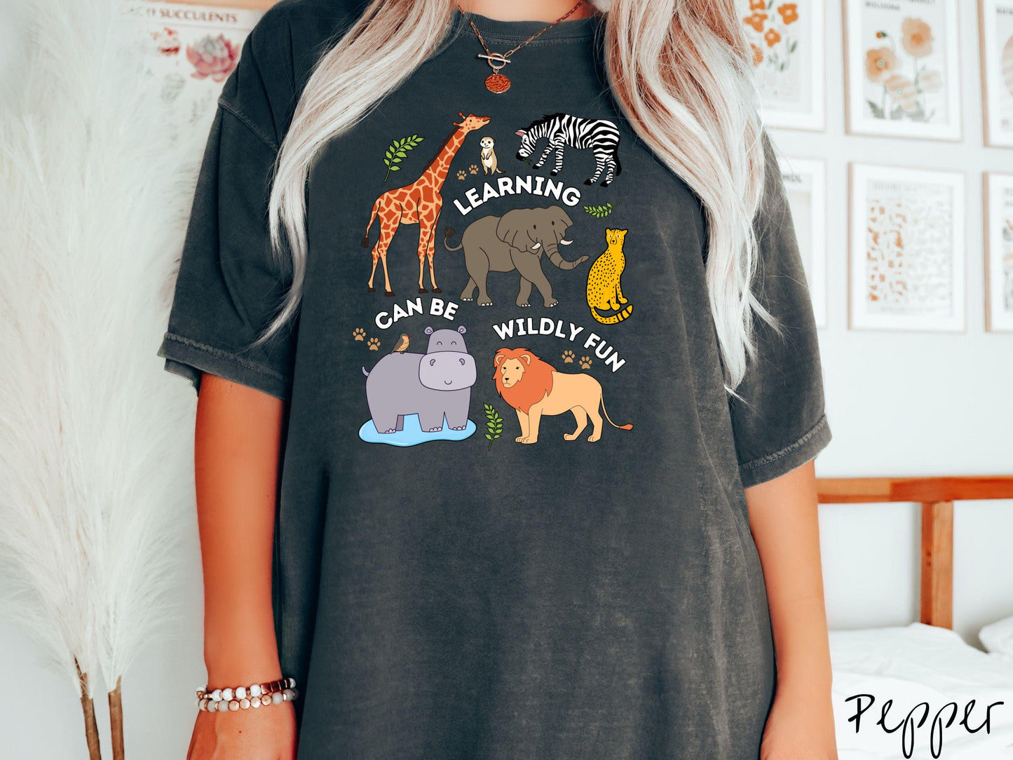 A woman wearing a cute, vintage pepper colored Comfort Colors T-shirt with the text Learning Can Be Wildly Fun. There is a giraffe, zebra elephant, hippopotamus, lion, leopard, and ground squirrel surrounded by leafy branches and paw prints.