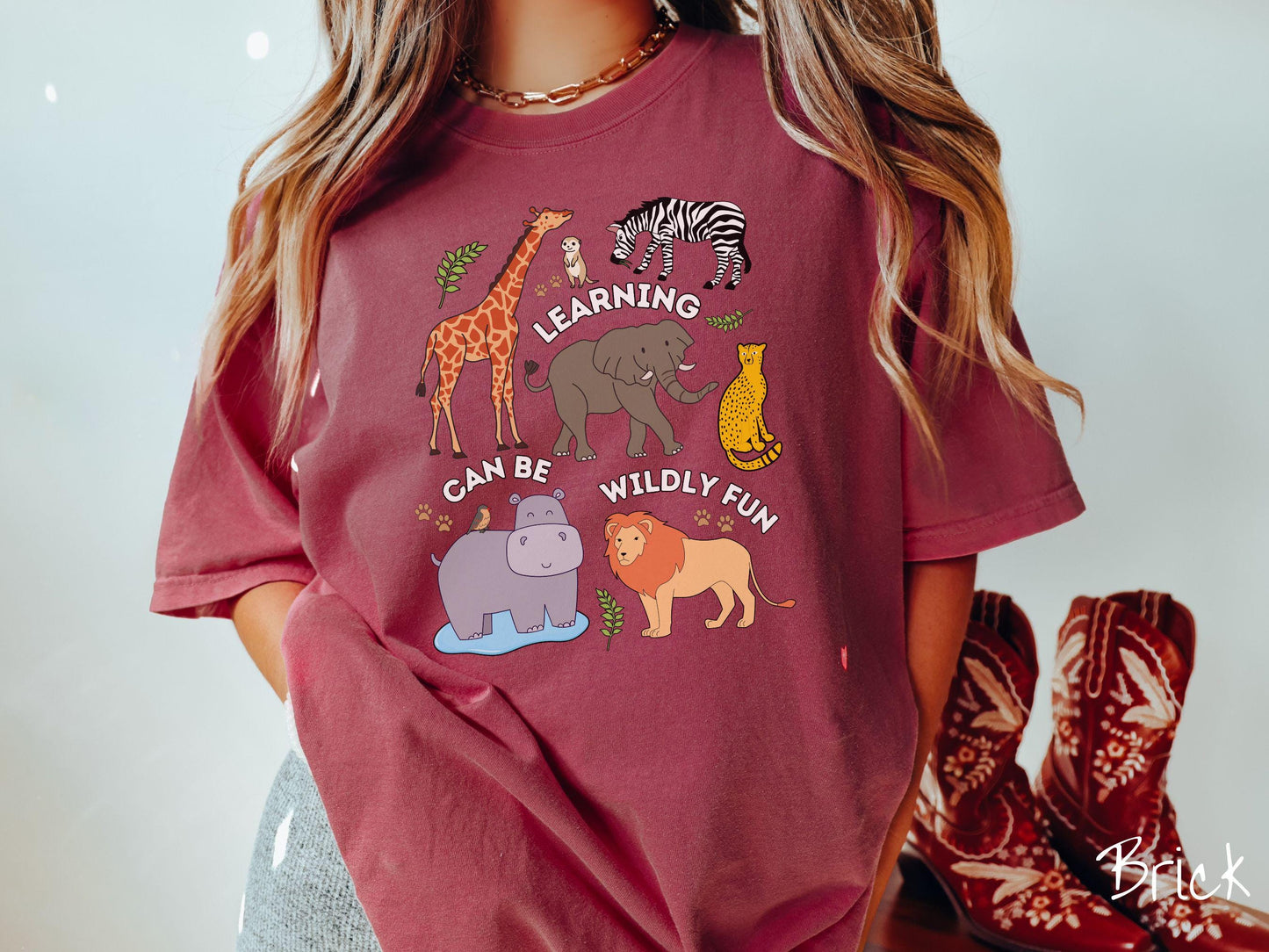 A woman wearing a cute, vintage brick colored Comfort Colors T-shirt with the text Learning Can Be Wildly Fun. There is a giraffe, zebra elephant, hippopotamus, lion, leopard, and ground squirrel surrounded by leafy branches and paw prints.