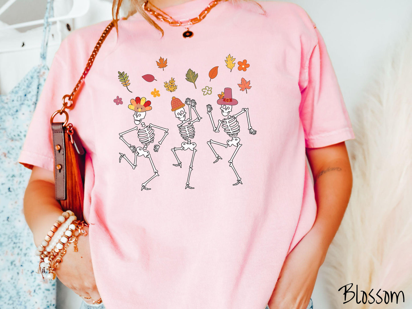A woman wearing a cute blossom colored Comfort Colors t-shirt with three white skeletons wearing pilgrim hats dancing with colorful fall leaves in the air.