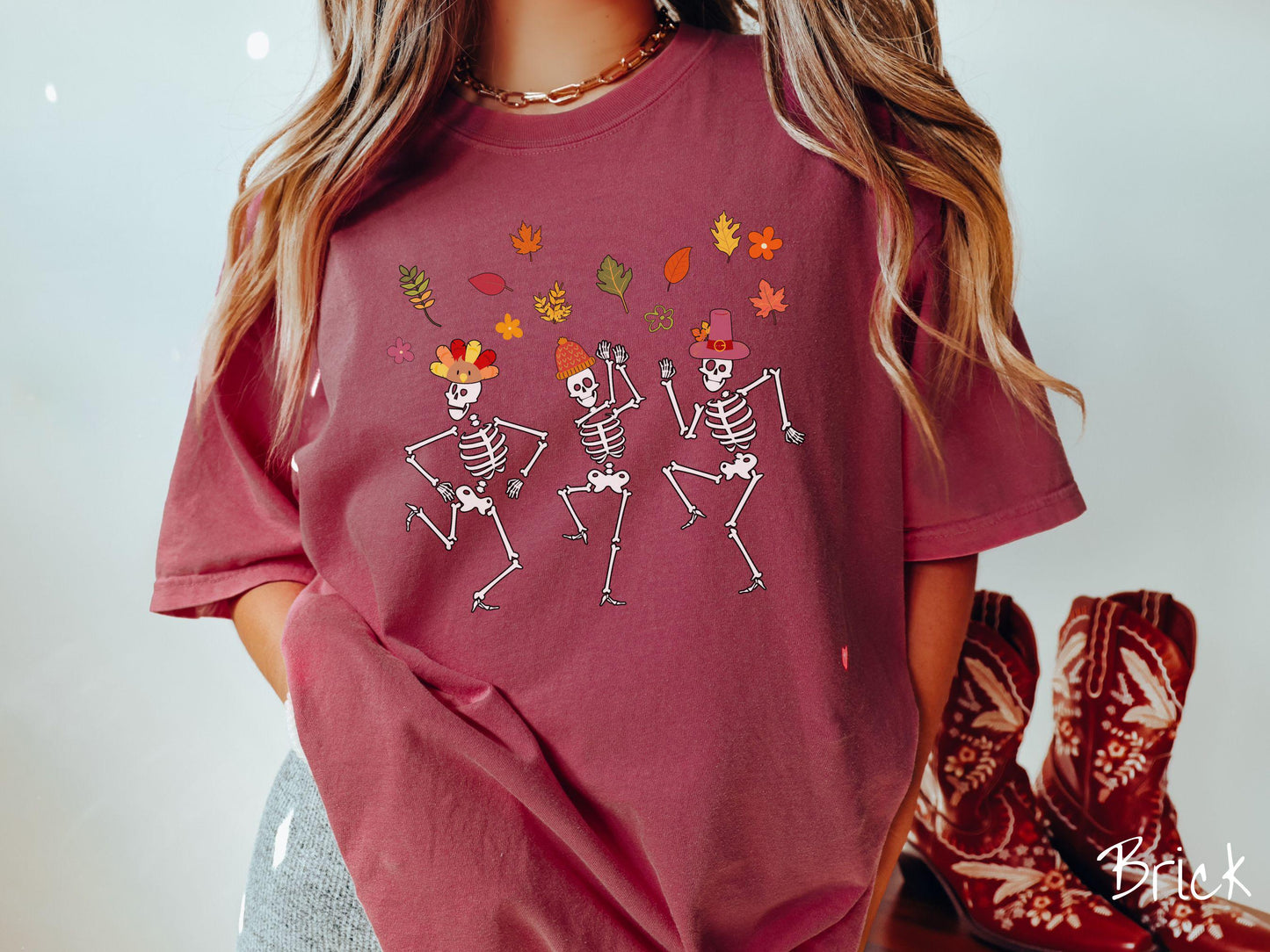 A woman wearing a cute brick colored Comfort Colors t-shirt with three white skeletons wearing pilgrim hats dancing with colorful fall leaves in the air.