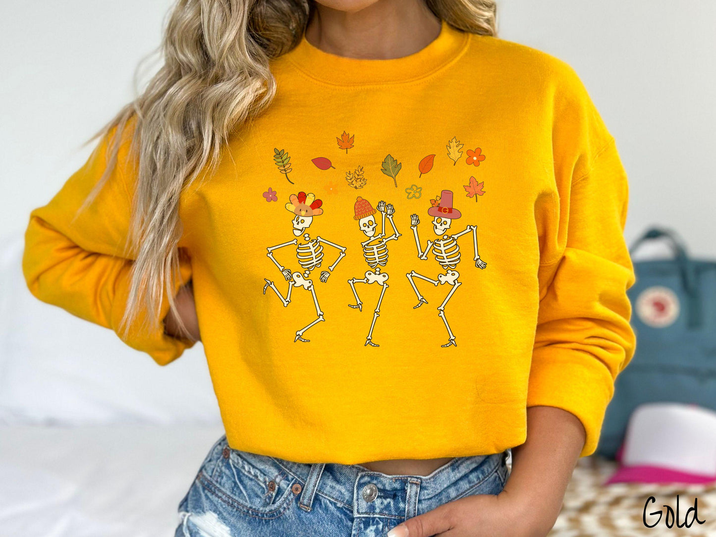 A woman wearing a cute gold colored sweatshirt with three white skeletons wearing pilgrim hats dancing with colorful fall leaves in the air.