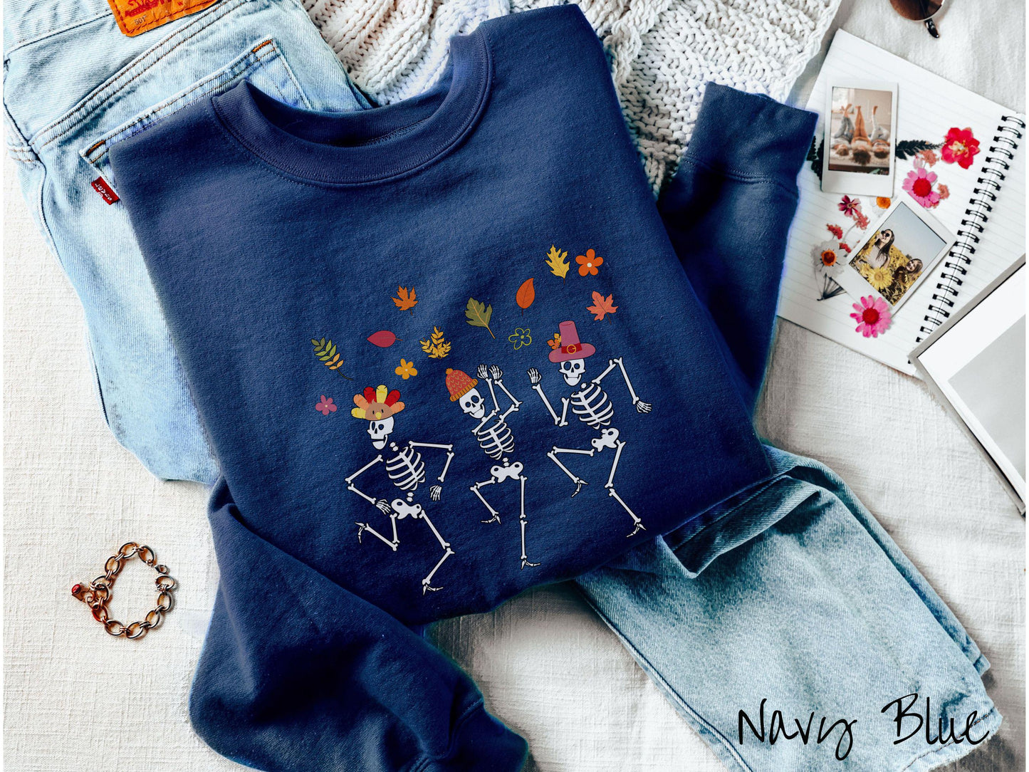 A woman wearing a cute navy colored sweatshirt with three white skeletons wearing pilgrim hats dancing with colorful fall leaves in the air.