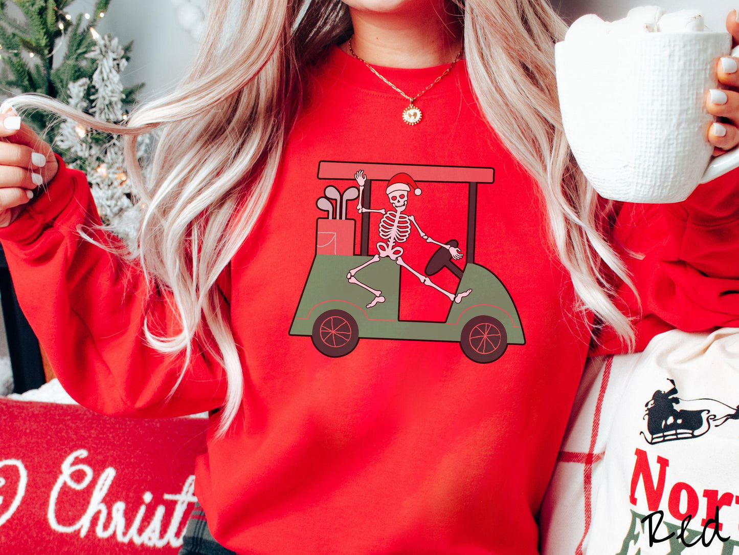 A woman wearing a cute red colored sweatshirt with smiling Skeleton with a Santa Hat driving a green golf cart and waving with one hand