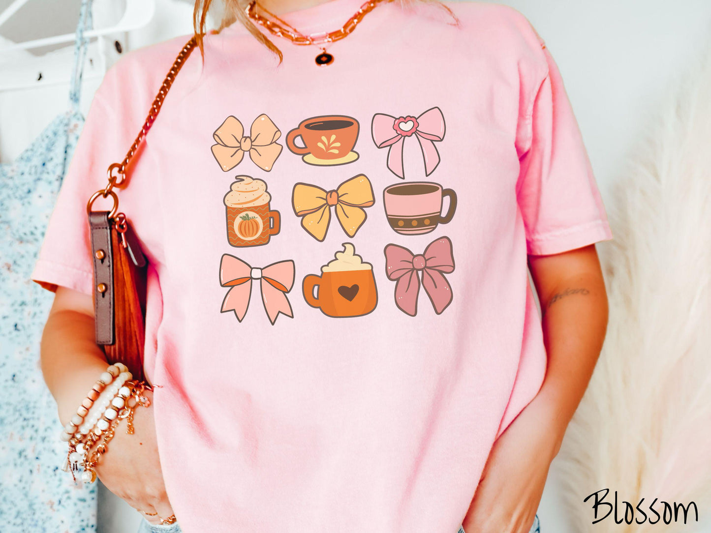 A woman wearing a cute, vintage blossom colored Comfort Colors T-shirt with a grid of pictures of an orange bow, an orange tea cup, a pink bow, an orange pumpkin-spiced latte, a yellow bow, a pink coffee cup, a pink bow, and an orange coffee drink.