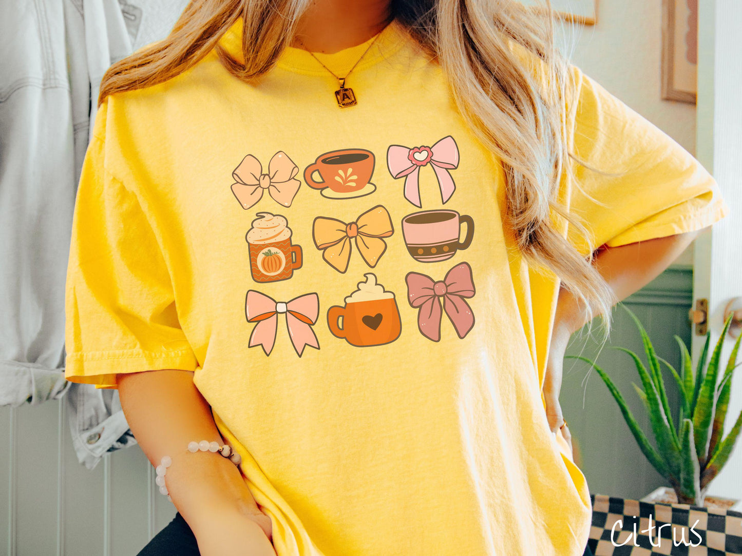 A woman wearing a cute, vintage citrus colored Comfort Colors T-shirt with a grid of pictures of an orange bow, an orange tea cup, a pink bow, an orange pumpkin-spiced latte, a yellow bow, a pink coffee cup, a pink bow, and an orange coffee drink.