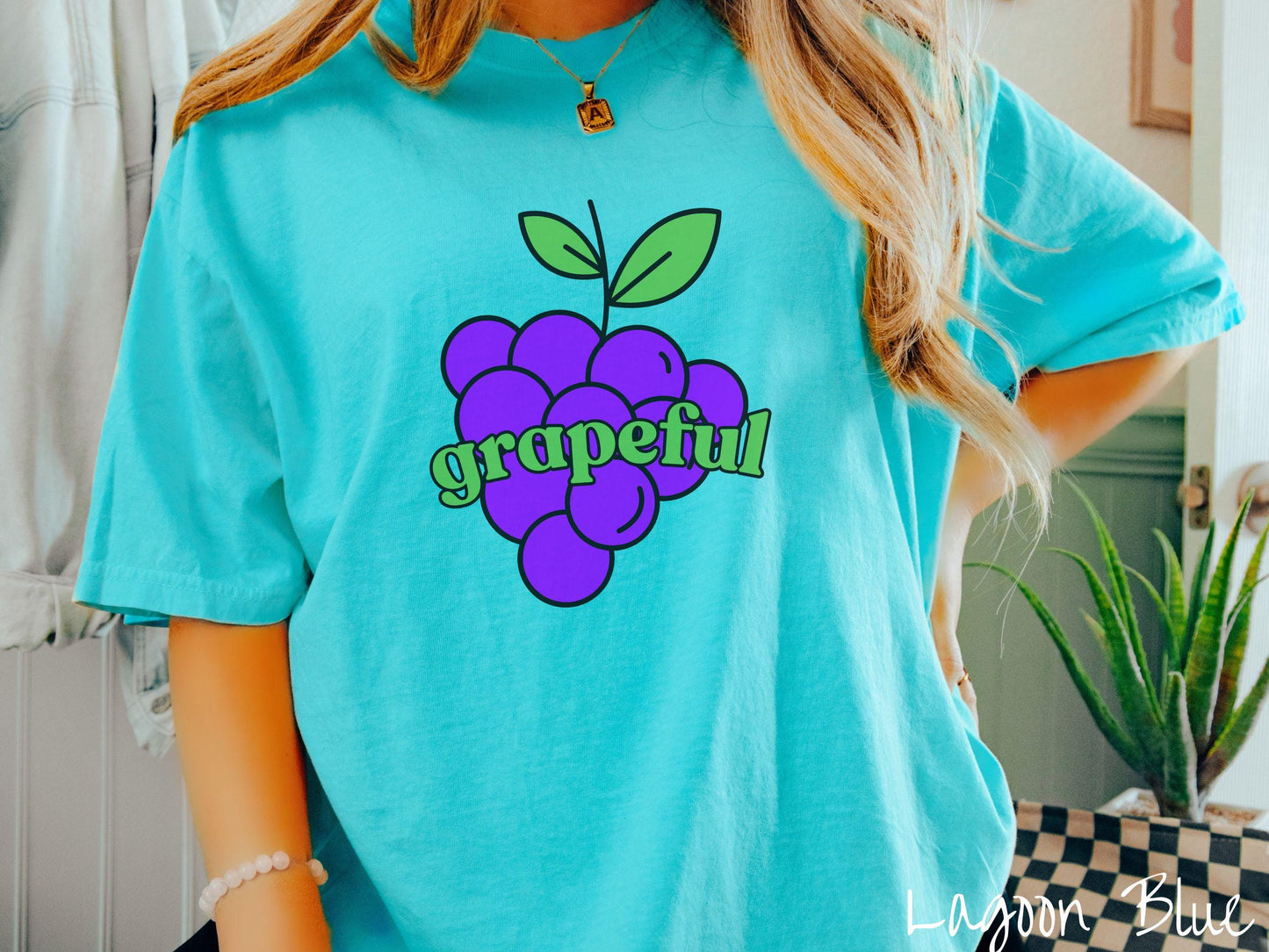 A woman wearing a cute, vintage lagoon blue colored Comfort Colors T-shirt with the text grapeful in green font across the front. Behind grapeful is a bunch of juicy, purple grapes with a green stem.