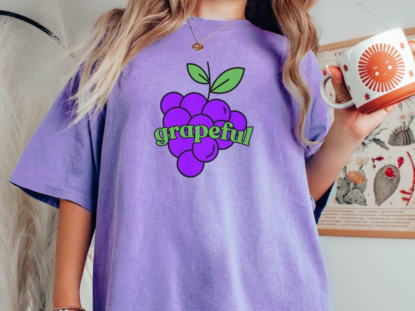 A woman wearing a cute, vintage violet colored Comfort Colors T-shirt with the text grapeful in green font across the front. Behind grapeful is a bunch of juicy, purple grapes with a green stem.