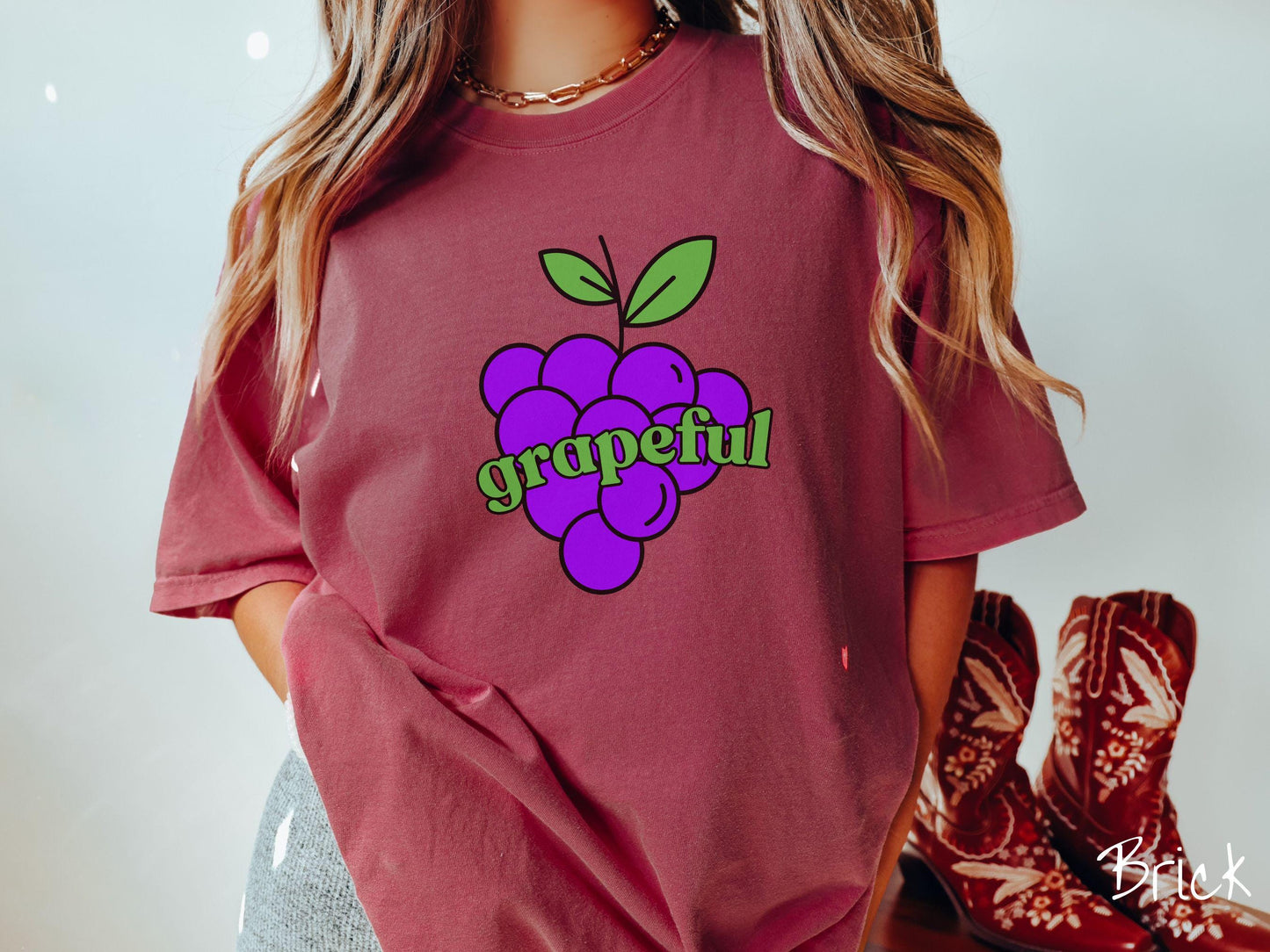 A woman wearing a cute, vintage brick colored Comfort Colors T-shirt with the text grapeful in green font across the front. Behind grapeful is a bunch of juicy, purple grapes with a green stem.