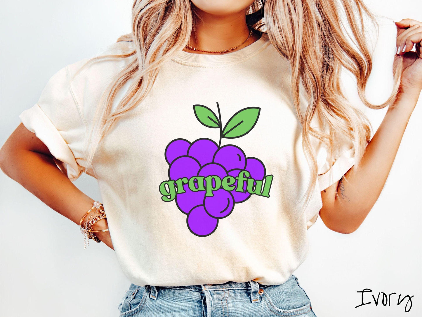 A woman wearing a cute, vintage ivory colored Comfort Colors T-shirt with the text grapeful in green font across the front. Behind grapeful is a bunch of juicy, purple grapes with a green stem.