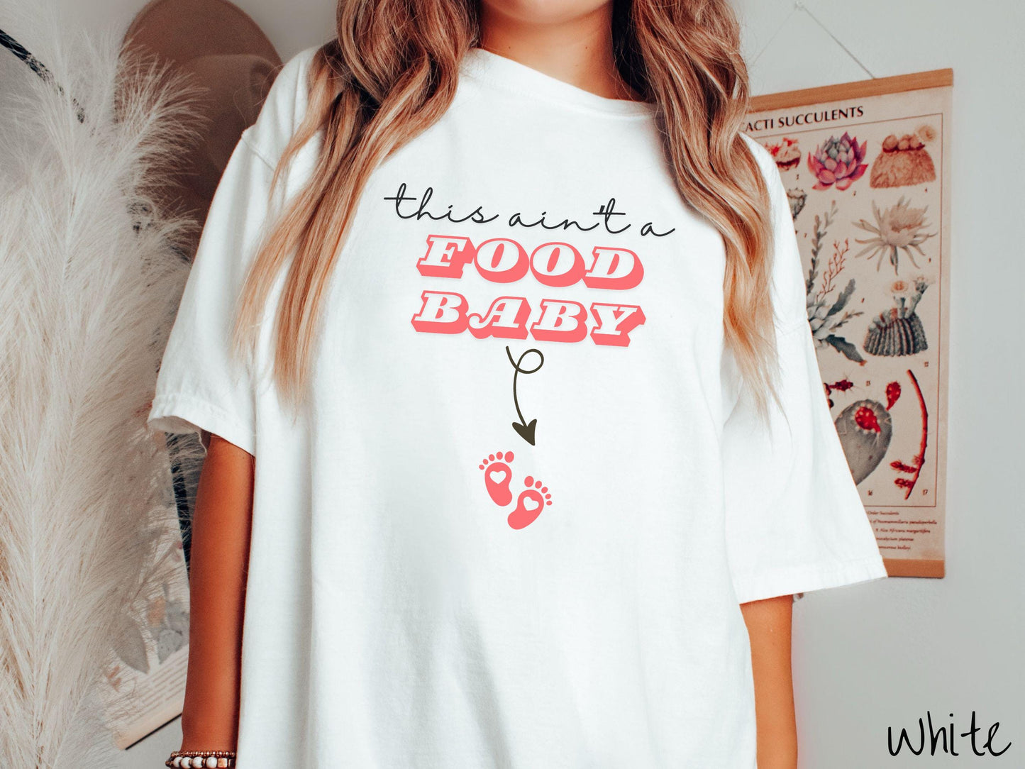 A woman wearing a cute, vintage white colored Comfort Colors T-shirt with the text this aint a food baby in black and red font. There is a black arrow pointing down to a pair of red baby footprints with hearts in them.