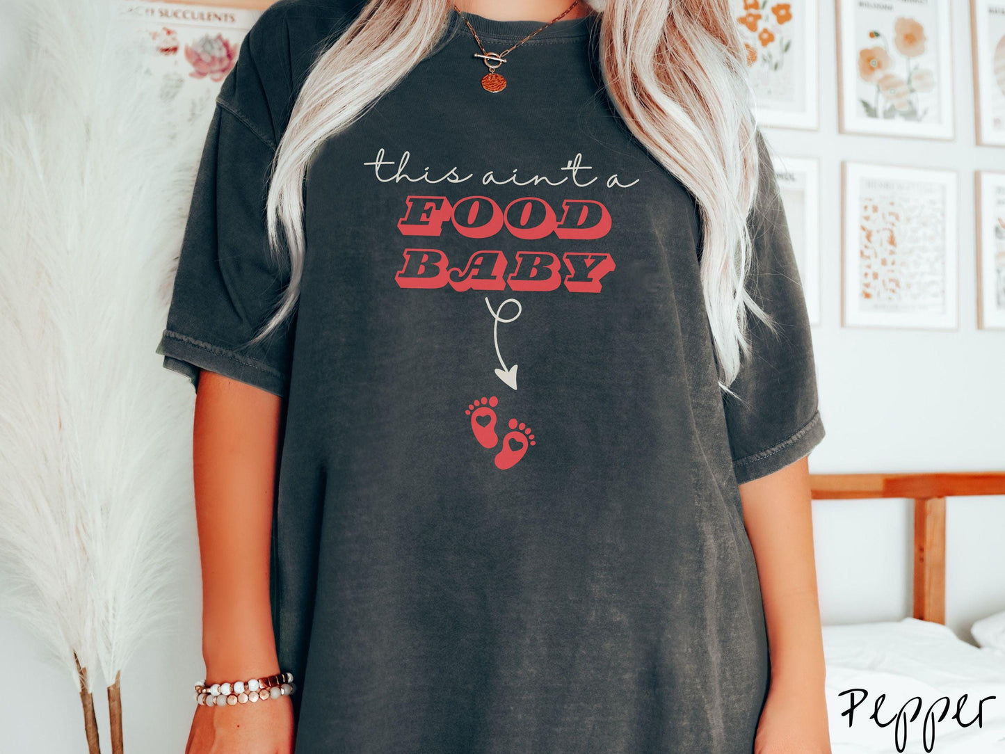 A woman wearing a cute, vintage pepper colored Comfort Colors T-shirt with the text this aint a food baby in black and red font. There is a black arrow pointing down to a pair of red baby footprints with hearts in them.