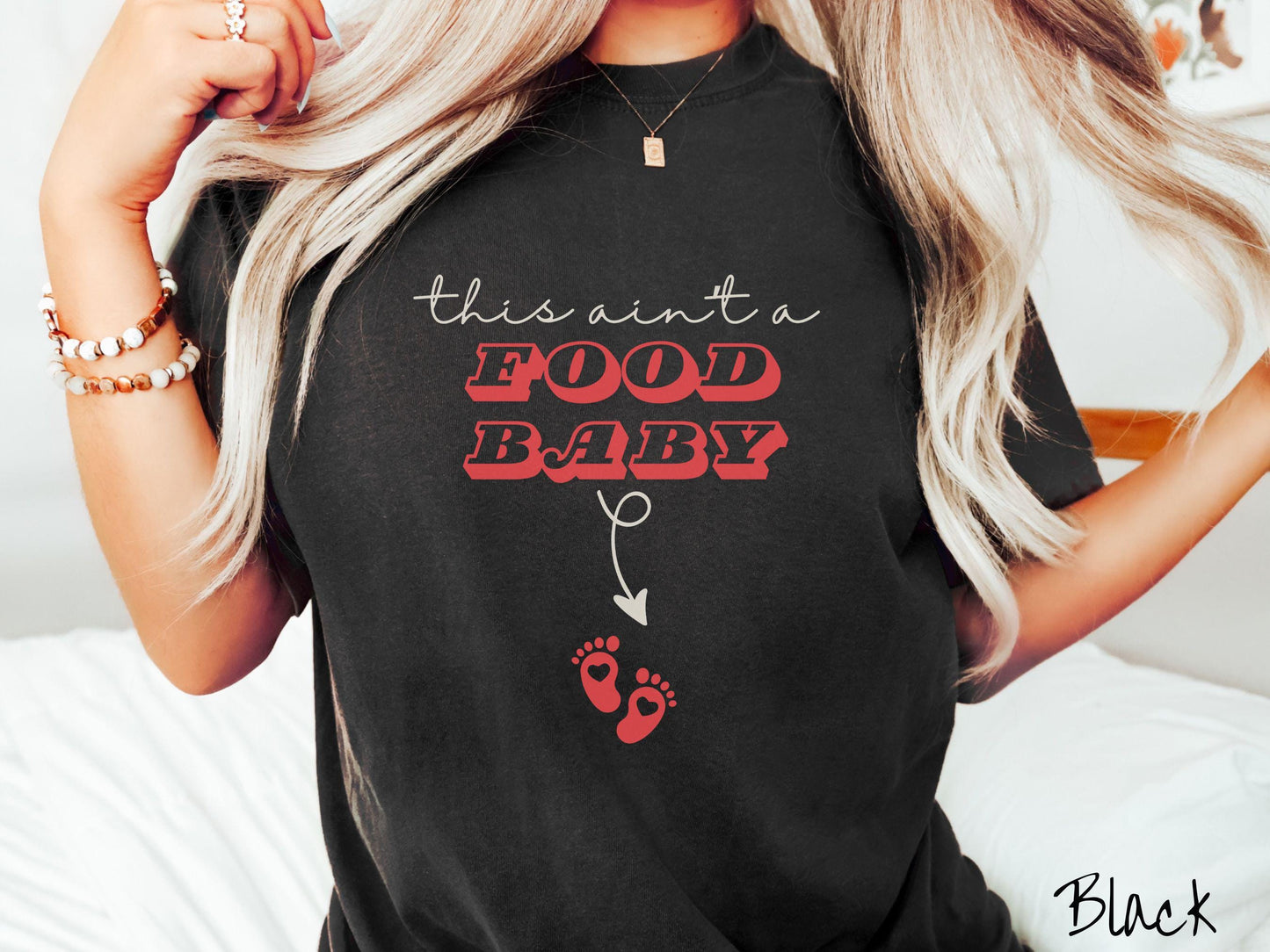 A woman wearing a cute, vintage black colored Comfort Colors T-shirt with the text this aint a food baby in black and red font. There is a black arrow pointing down to a pair of red baby footprints with hearts in them.