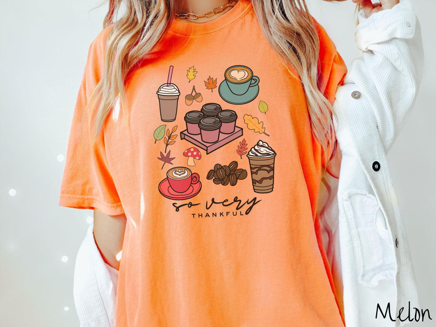 A woman wearing a cute, vintage melon colored Comfort Colors T-shirt with the text so very thankful. Above are an iced coffee, four to-go cup coffees, a latte, a frappucino, coffee beans, mushrooms, and fall leaves scattered around.