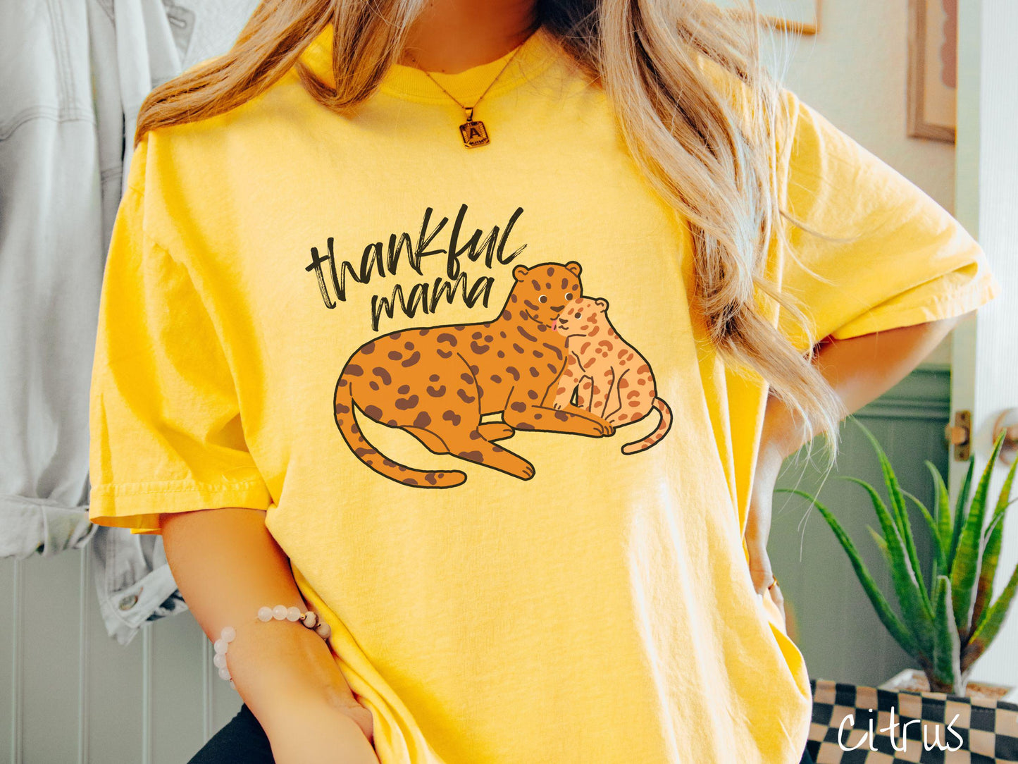 A woman wearing a cute, vintage citrus colored Comfort Colors T-shirt with the text thankful mama in black, sharpie font. Below is an orange, mother leopard coddling its child.