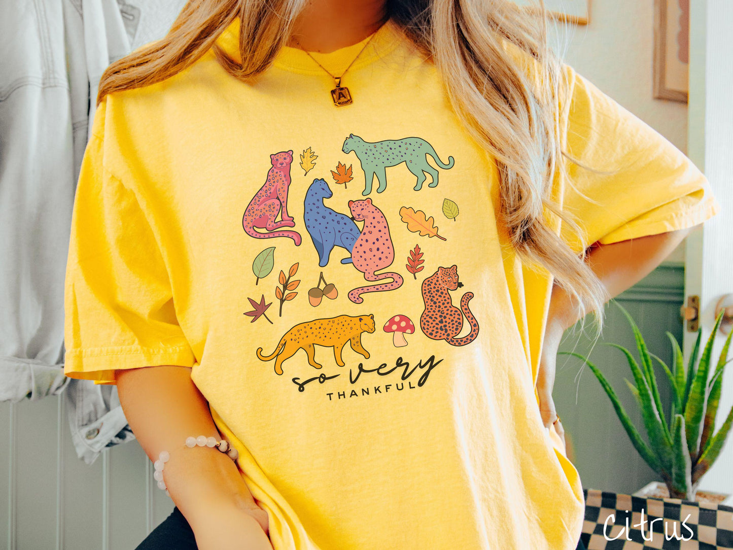 A woman wearing a cute, vintage citrus colored Comfort Colors T-shirt with the text so very thankful in black font. Above are colorful, spotted leopards in seated and walking positions, as well as leaves, acorns, and mushrooms scattered about.