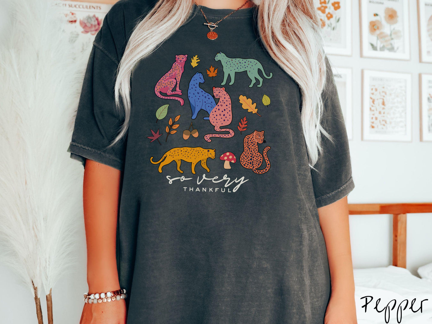A woman wearing a cute, vintage pepper colored Comfort Colors T-shirt with the text so very thankful in black font. Above are colorful, spotted leopards in seated and walking positions, as well as leaves, acorns, and mushrooms scattered about.