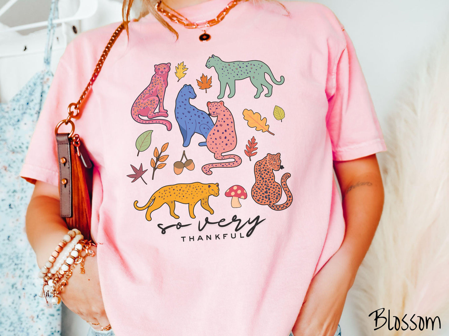 A woman wearing a cute, vintage blossom colored Comfort Colors T-shirt with the text so very thankful in black font. Above are colorful, spotted leopards in seated and walking positions, as well as leaves, acorns, and mushrooms scattered about.