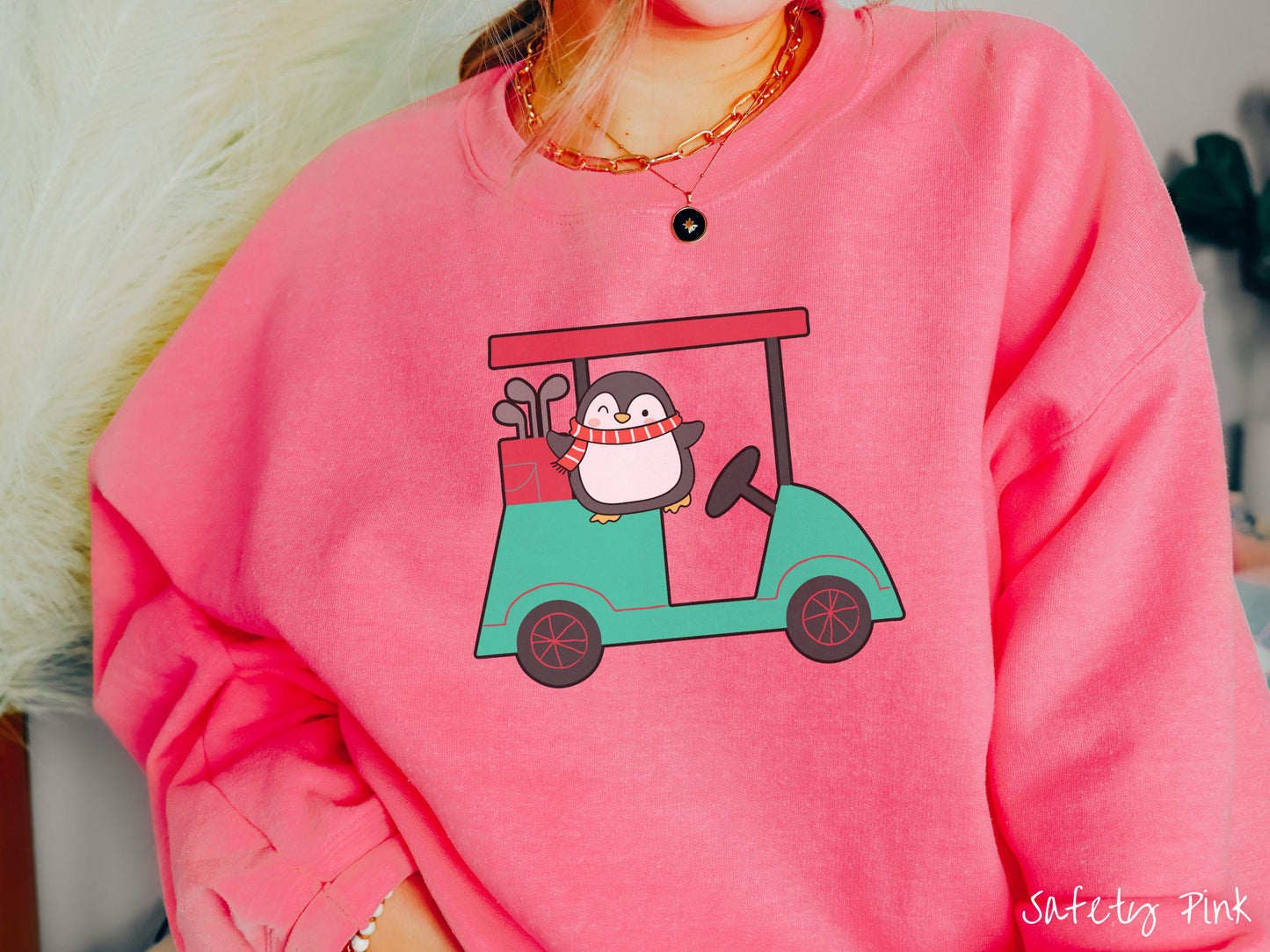 A woman wearing a cute safety pink colored sweatshirt with smiling penguin driving a green golf cart and waving with both hands.
