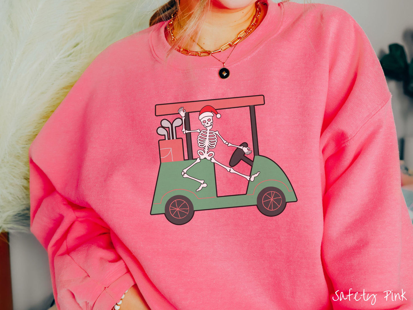 A woman wearing a cute safety pink colored sweatshirt with smiling Skeleton with a Santa Hat driving a green golf cart and waving with one hand