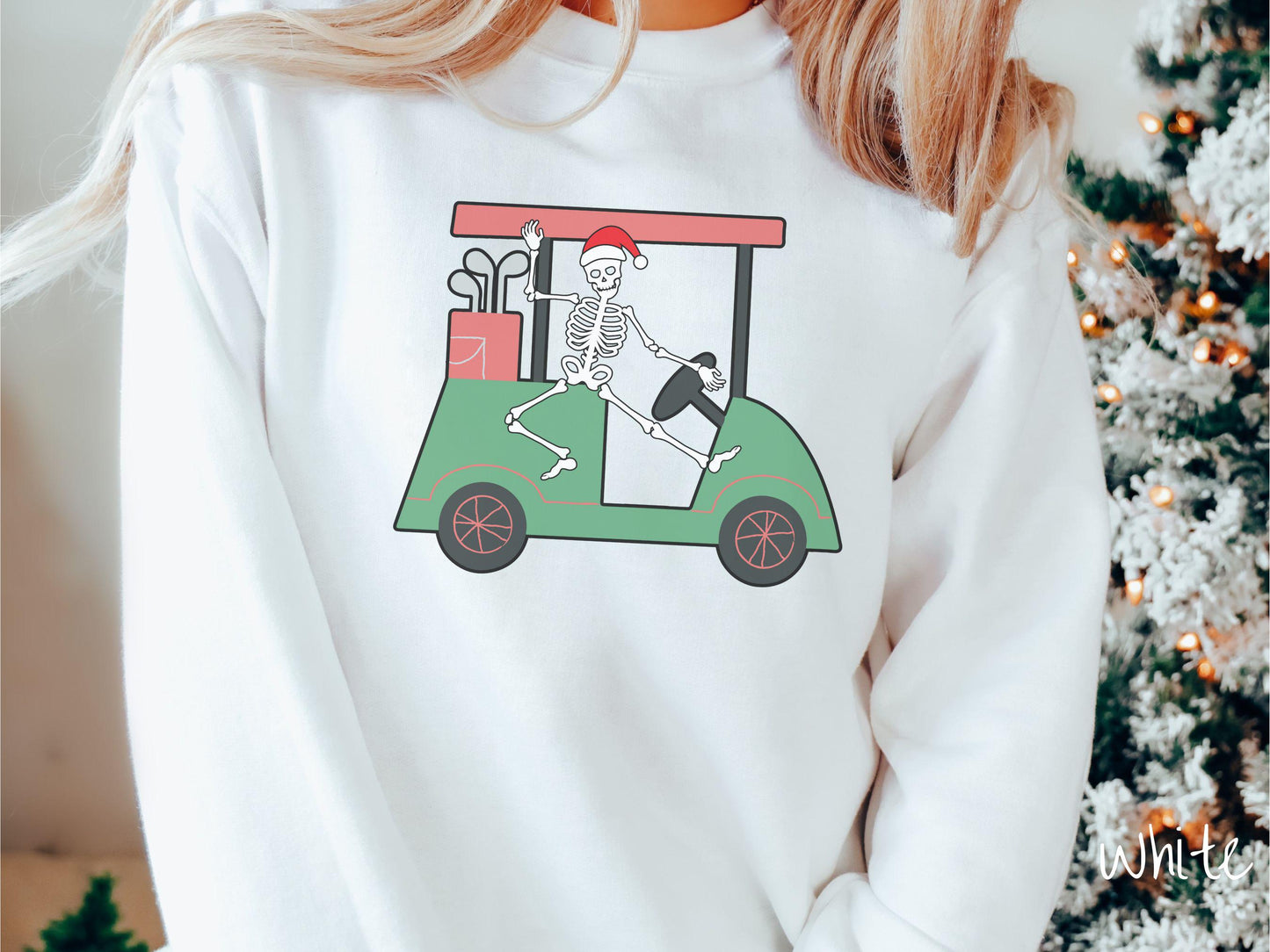 A woman wearing a cute white colored sweatshirt with smiling Skeleton with a Santa Hat driving a green golf cart and waving with one hand