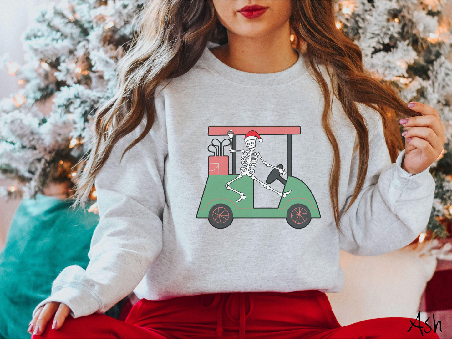 A woman wearing a cute ash colored sweatshirt with smiling Skeleton with a Santa Hat driving a green golf cart and waving with one hand