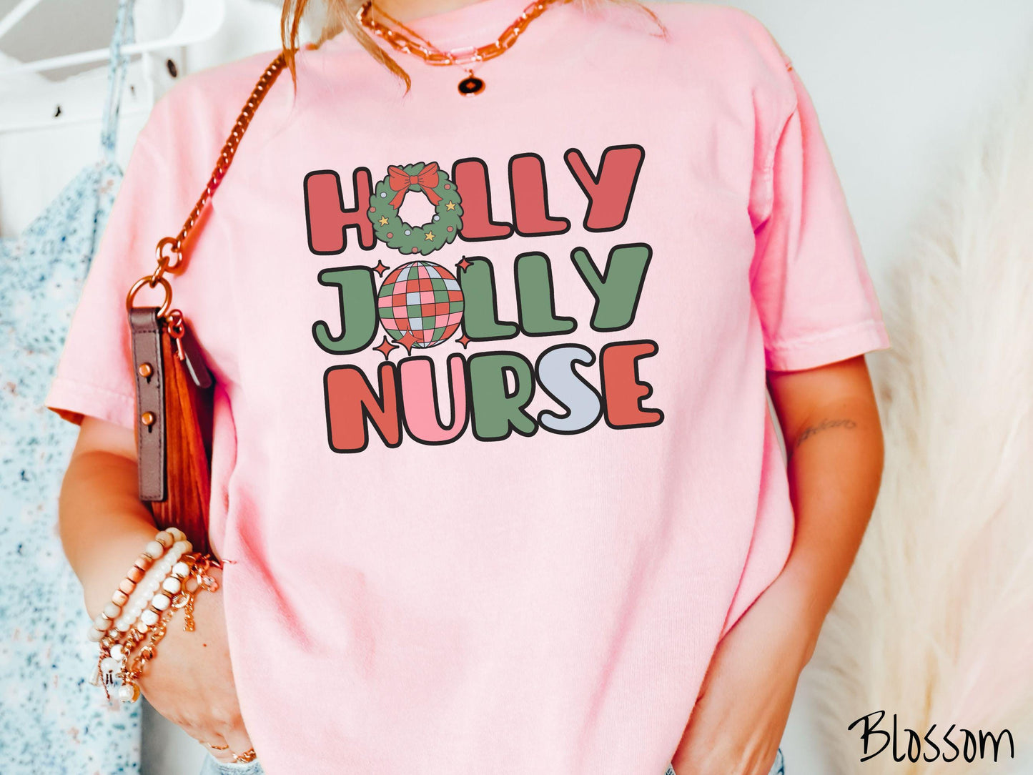 A woman wearing a cute, vintage blossom colored Comfort Colors T-shirt with text Holly Jolly Nurse in red, green , pink, and cadet blue font. The letter O in Holly is a Christmas wreath and the letter O in Jolly is a red, green, and blue disco ball.