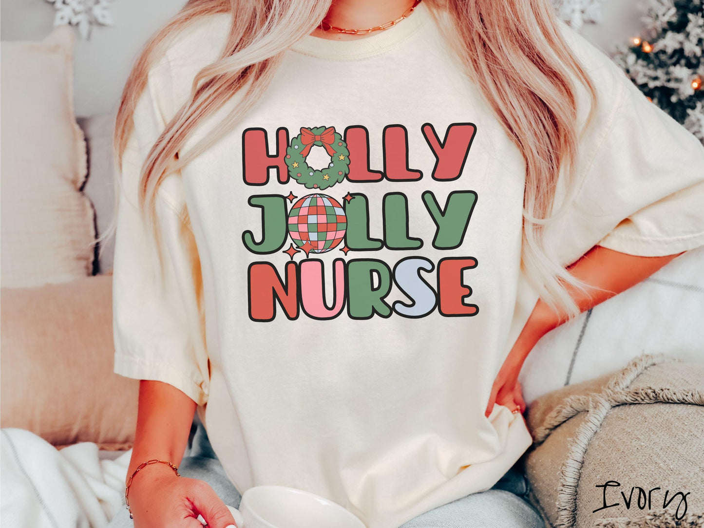 A woman wearing a cute, vintage ivory colored Comfort Colors T-shirt with text Holly Jolly Nurse in red, green , pink, and cadet blue font. The letter O in Holly is a Christmas wreath and the letter O in Jolly is a red, green, and blue disco ball.