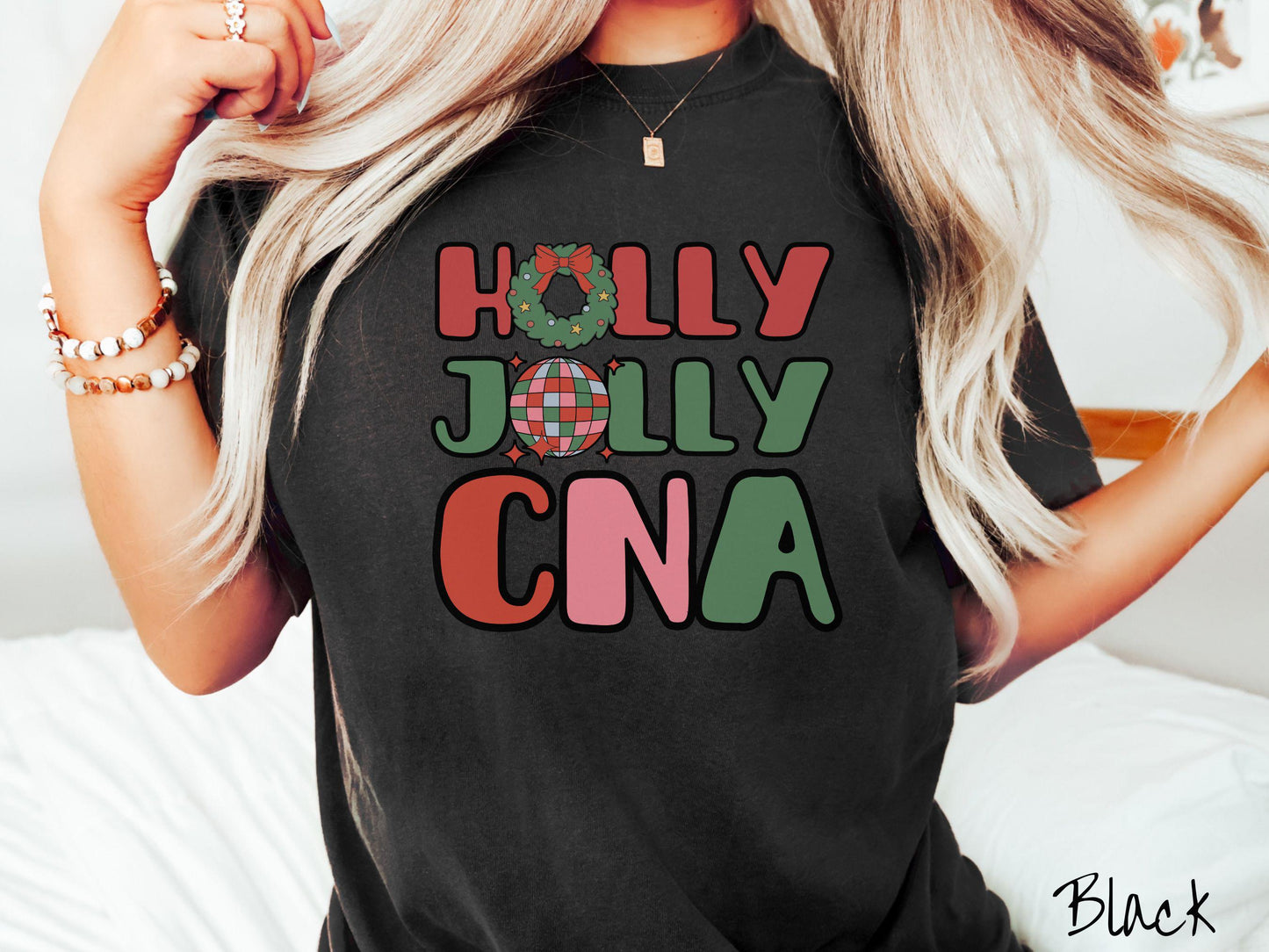 A woman wearing a cute, vintage black colored Comfort Colors T-shirt with the text Holly Jolly CNA in red, green, pink, and blue font. The letter O in Holly is a Christmas wreath and the letter O in Jolly is a multi-colored disco ball.