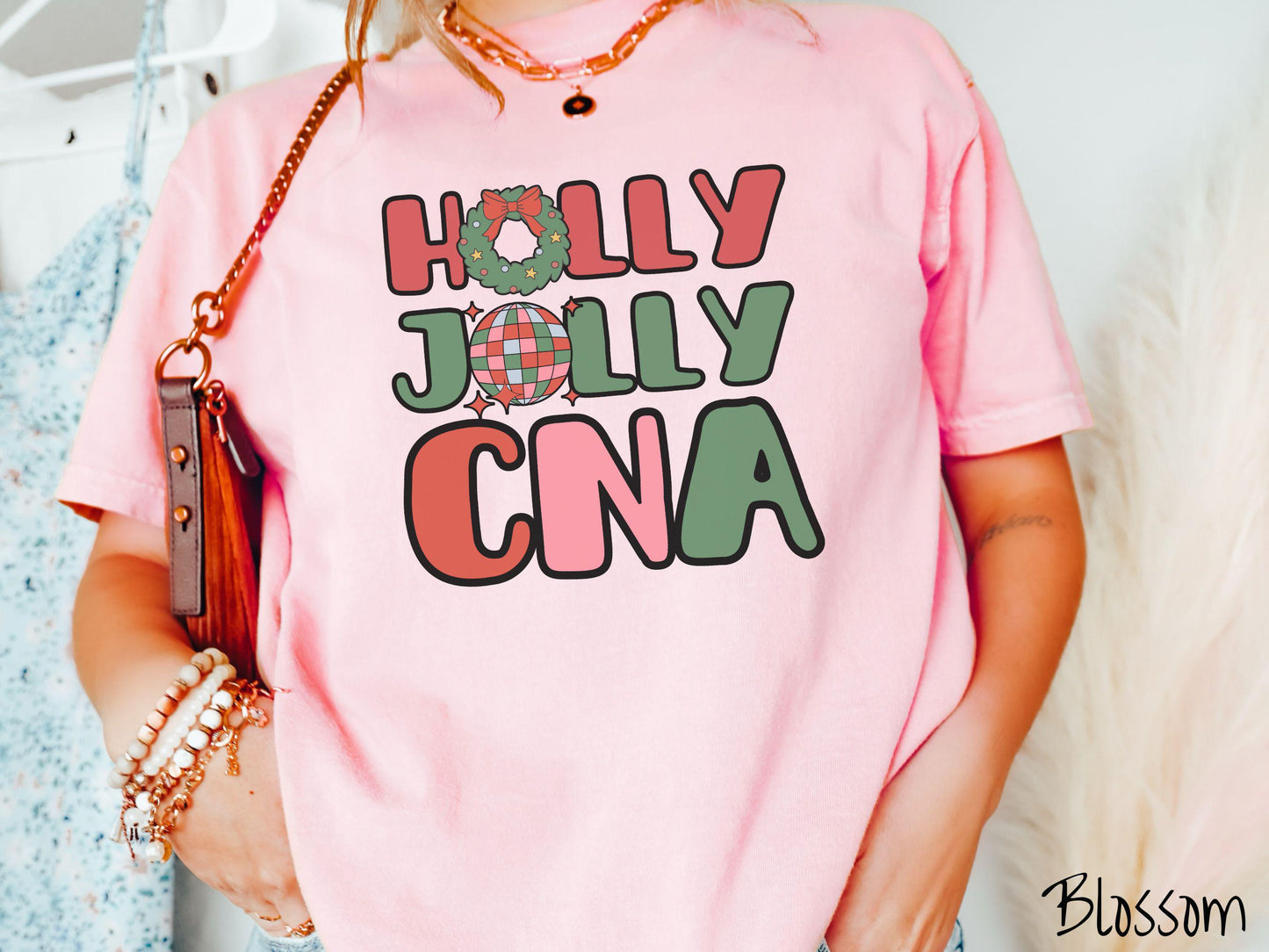 A woman wearing a cute, vintage blossom colored Comfort Colors T-shirt with the text Holly Jolly CNA in red, green, pink, and blue font. The letter O in Holly is a Christmas wreath and the letter O in Jolly is a multi-colored disco ball.