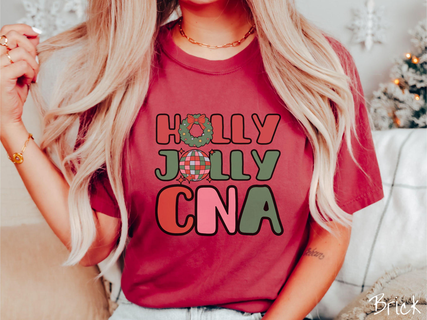 A woman wearing a cute, vintage brick colored Comfort Colors T-shirt with the text Holly Jolly CNA in red, green, pink, and blue font. The letter O in Holly is a Christmas wreath and the letter O in Jolly is a multi-colored disco ball.