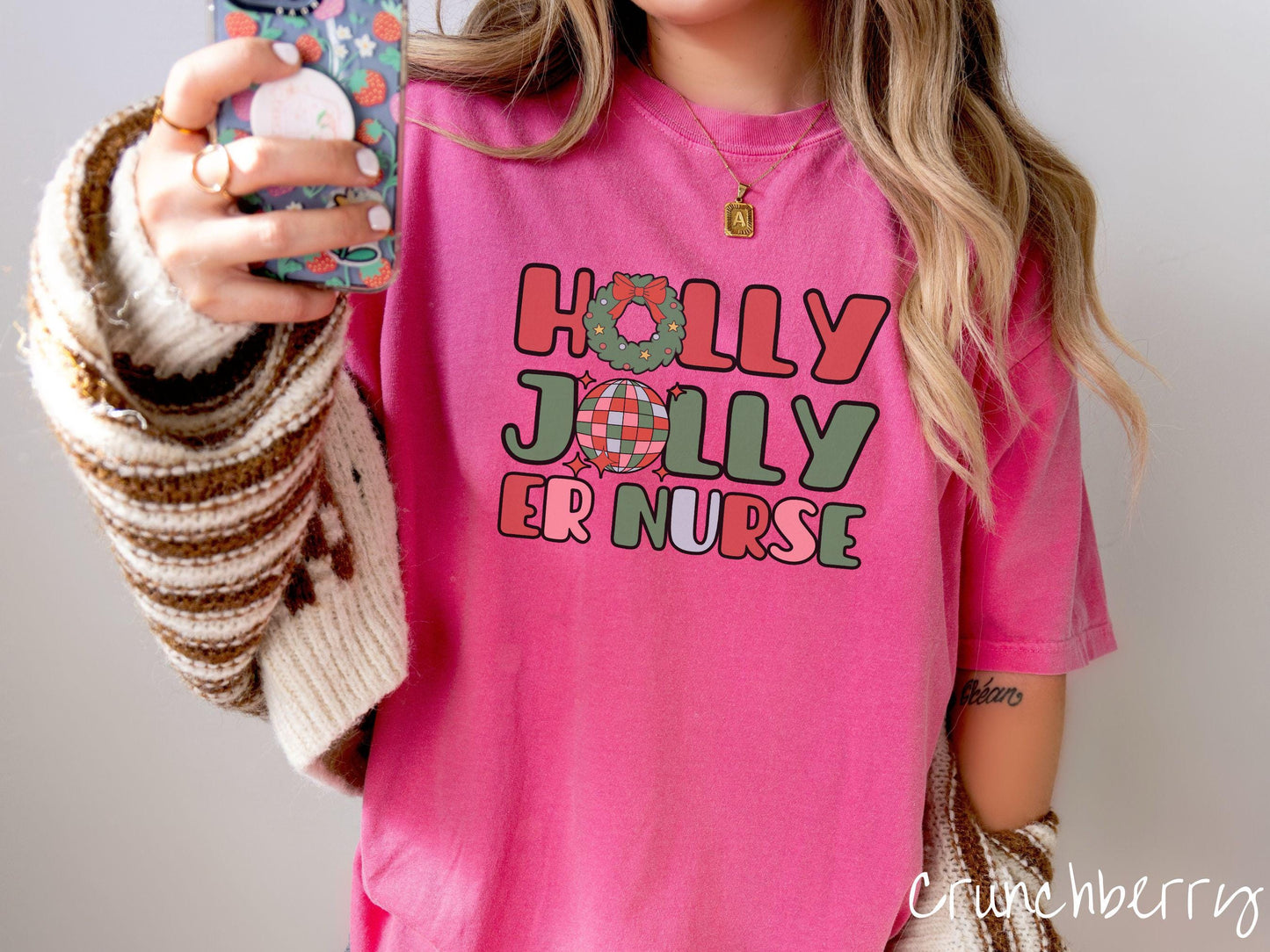 A woman wearing a cute, vintage crunchberry colored Comfort Colors T-shirt with the text Holly Jolly ER Nurse in red, green, pink, and blue font. The letter O in Holly is a Christmas wreath and the letter O in Jolly is a multi-colored disco ball.