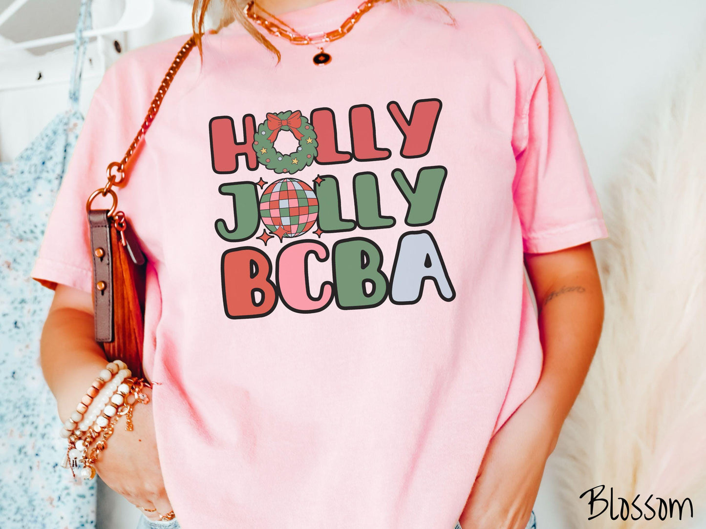 A woman wearing a cute, vintage blossom colored Comfort Colors T-shirt with the text Holly Jolly BCBA in red, green, pink, and blue font. The letter O in Holly is a Christmas wreath and the letter O in Jolly is a multi-colored disco ball.