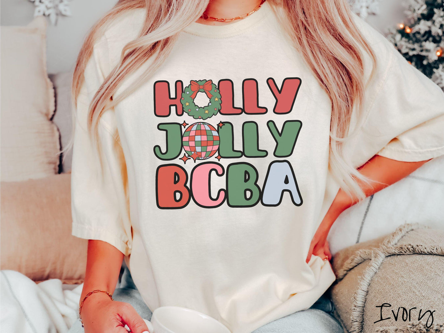 A woman wearing a cute, vintage ivory colored Comfort Colors T-shirt with the text Holly Jolly BCBA in red, green, pink, and blue font. The letter O in Holly is a Christmas wreath and the letter O in Jolly is a multi-colored disco ball.