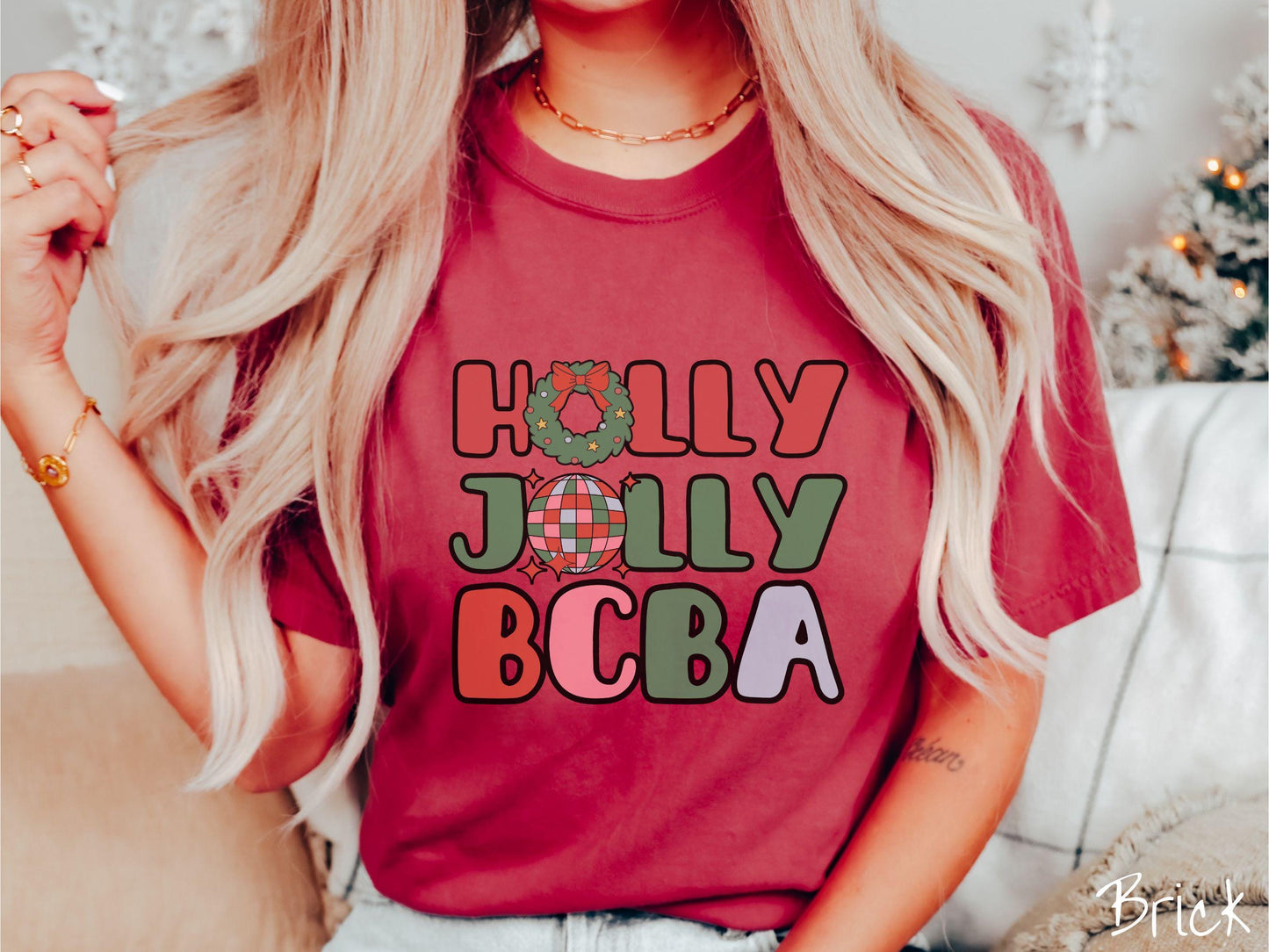 A woman wearing a cute, vintage brick colored Comfort Colors T-shirt with the text Holly Jolly BCBA in red, green, pink, and blue font. The letter O in Holly is a Christmas wreath and the letter O in Jolly is a multi-colored disco ball.