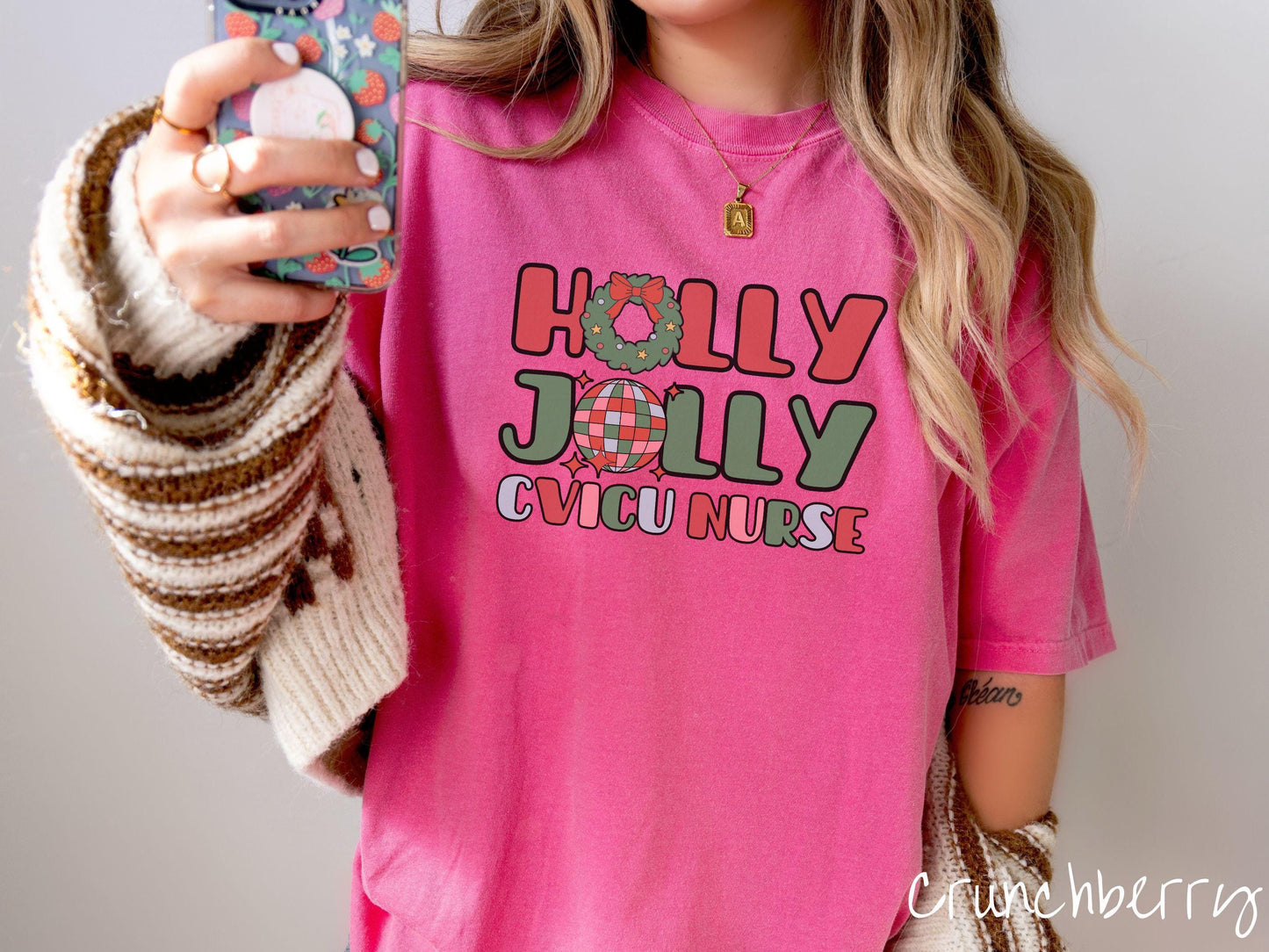 A woman wearing a cute, vintage crunchberry colored Comfort Colors T-shirt with the text Holly Jolly CVICU Nurse in red, green, pink, and blue font. The letter O in Holly is a Christmas wreath and the letter O in Jolly is a multi-colored disco ball.