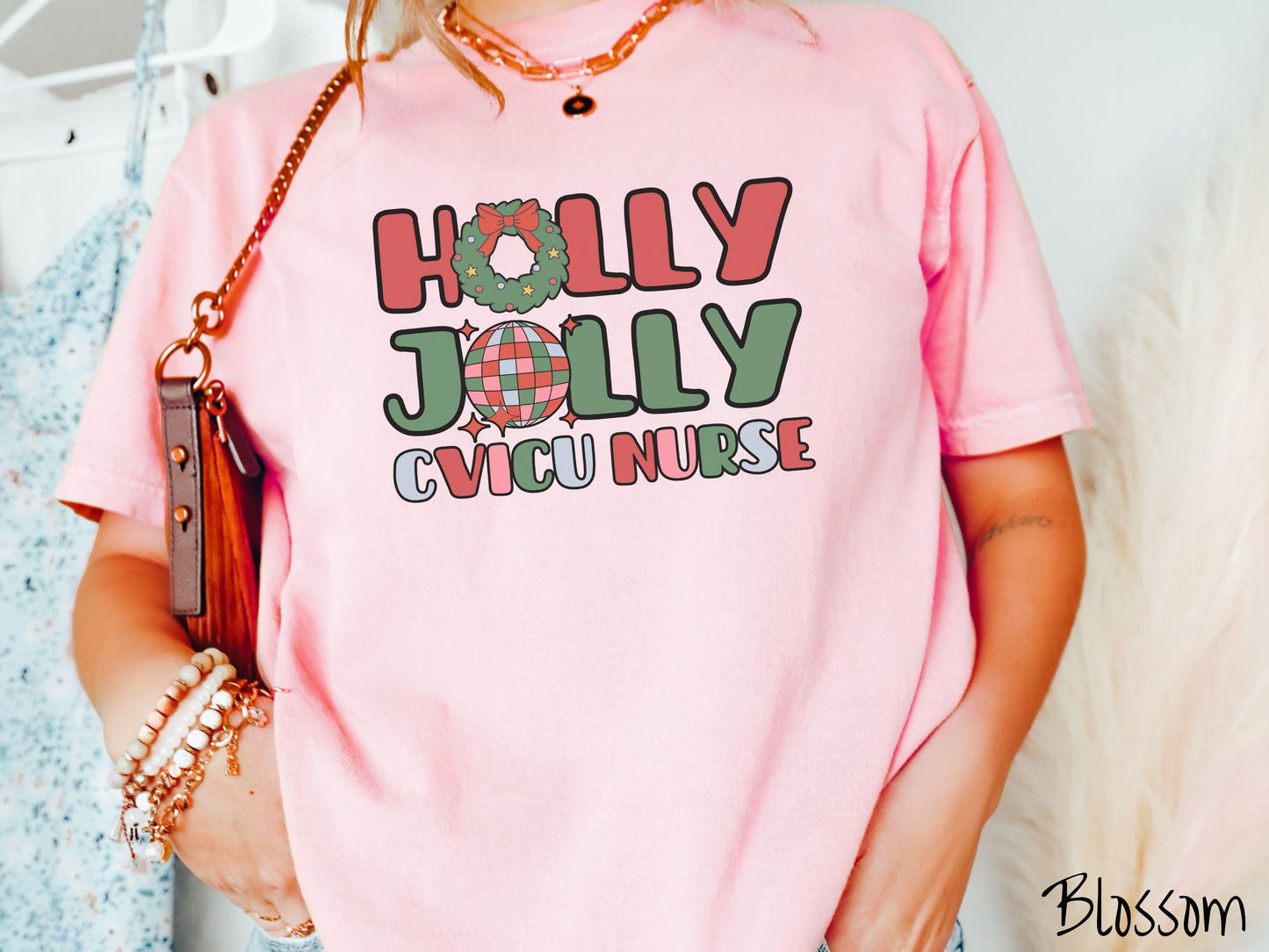 A woman wearing a cute, vintage blossom colored Comfort Colors T-shirt with the text Holly Jolly CVICU Nurse in red, green, pink, and blue font. The letter O in Holly is a Christmas wreath and the letter O in Jolly is a multi-colored disco ball.