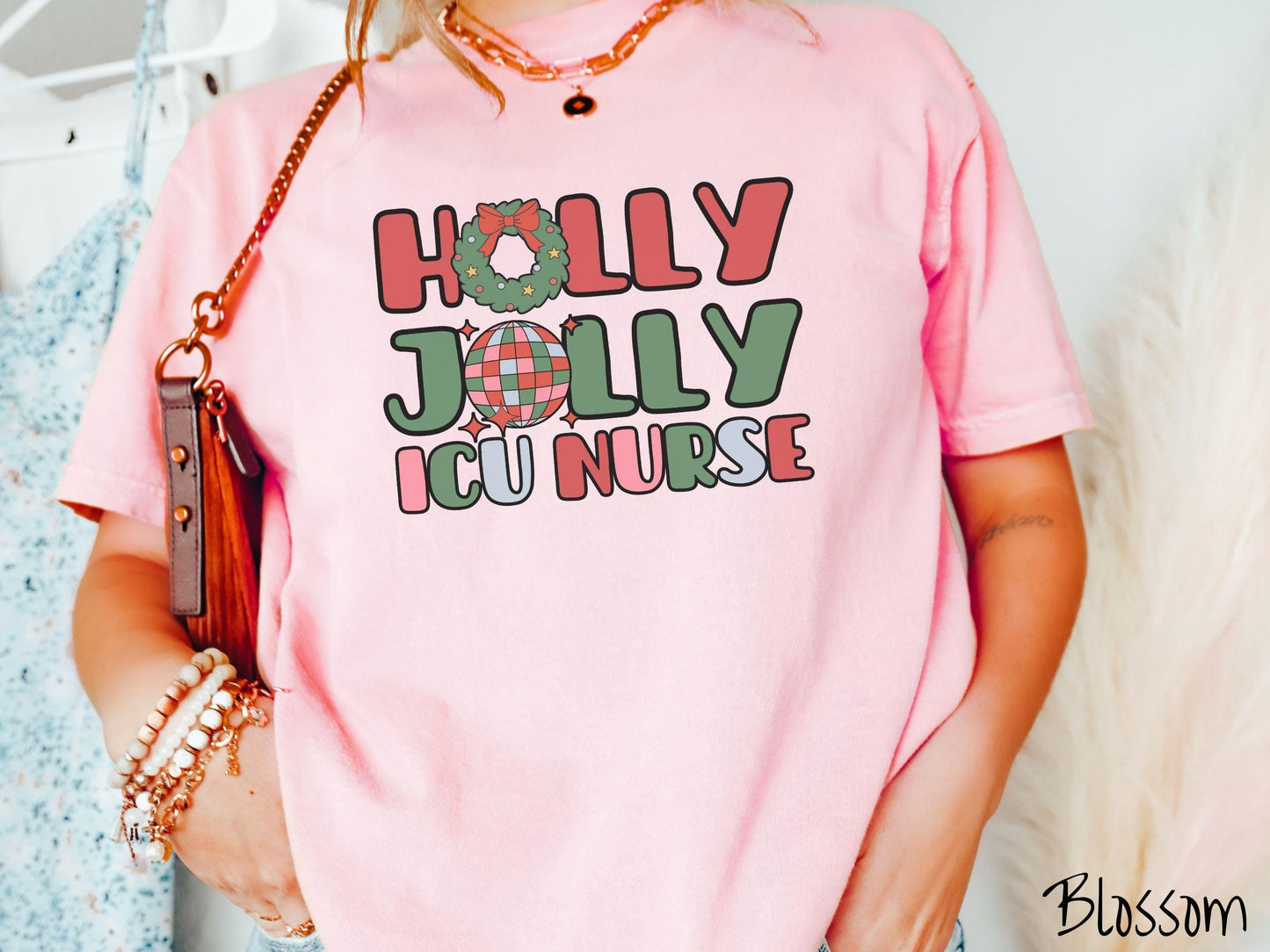 A woman wearing a cute, vintage blossom colored Comfort Colors T-shirt with the text Holly Jolly ICU Nurse in red, green, pink, and blue font. The letter O in Holly is a Christmas wreath and the letter O in Jolly is a multi-colored disco ball.