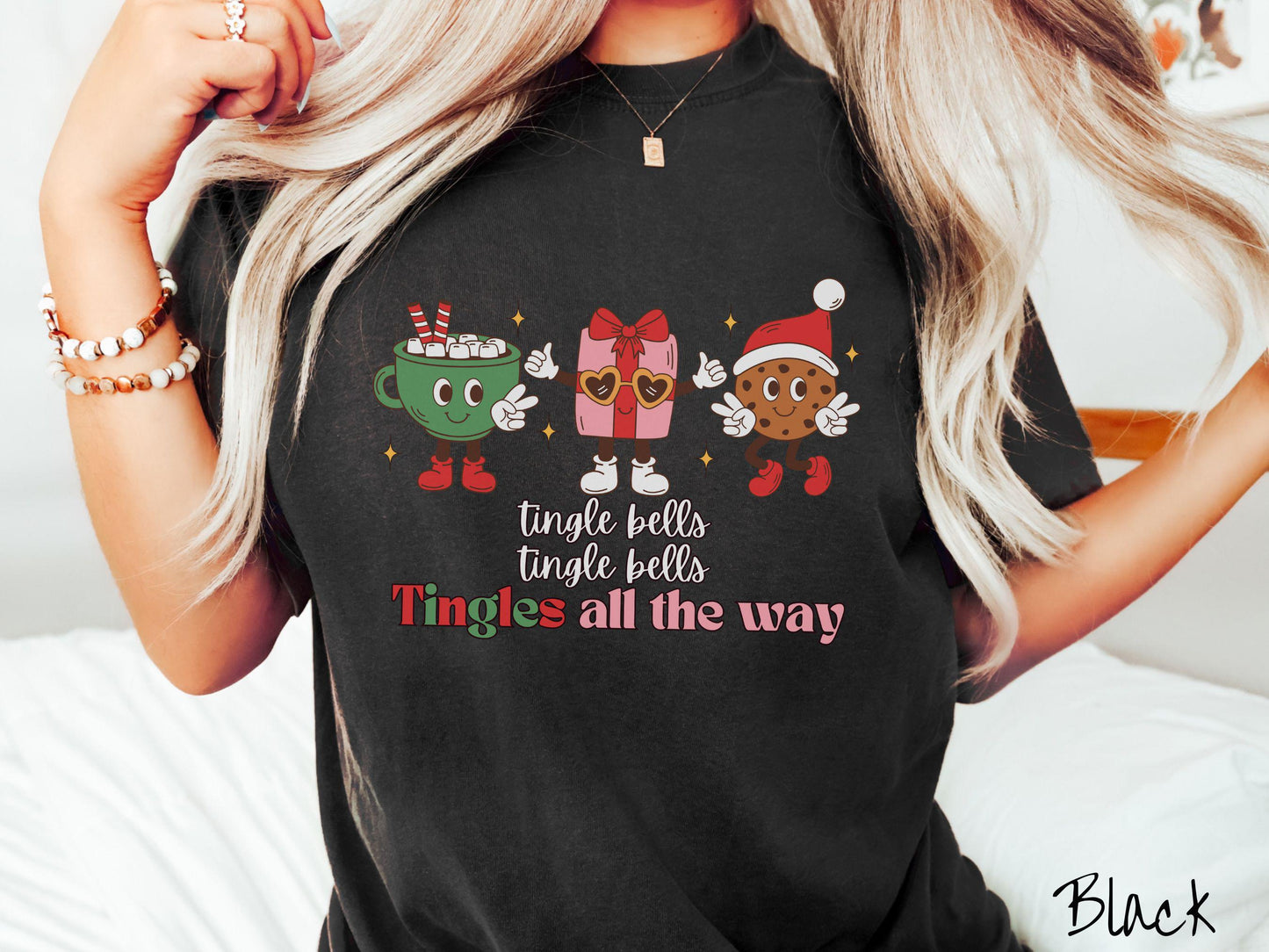 A cute, vintage black colored Comfort Colors T-shirt with the text tnigle bells, tingle bells, tingles all the way. Above are a green cup of hot cocoa holding a peace sign, a red present holding two thumbs up, and a cookie holding up peace signs.