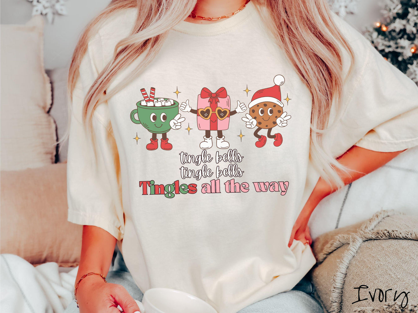 A cute, vintage ivory colored Comfort Colors T-shirt with the text tnigle bells, tingle bells, tingles all the way. Above are a green cup of hot cocoa holding a peace sign, a red present holding two thumbs up, and a cookie holding up peace signs.