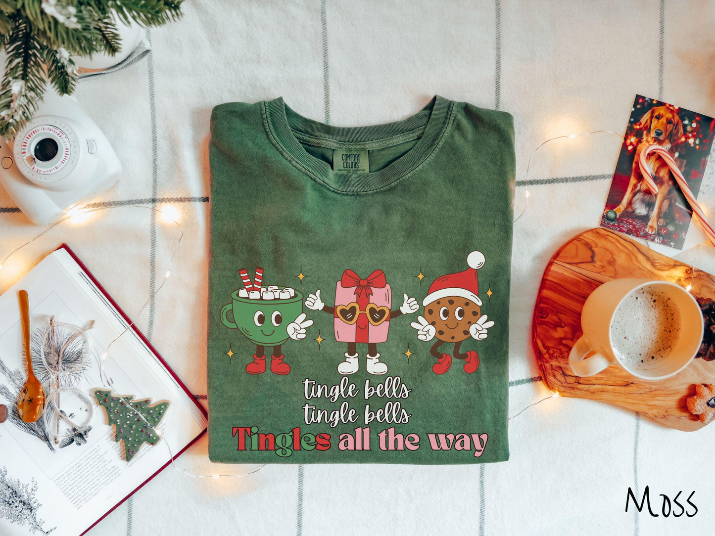 A cute, vintage moss colored Comfort Colors T-shirt with the text tnigle bells, tingle bells, tingles all the way. Above are a green cup of hot cocoa holding a peace sign, a red present holding two thumbs up, and a cookie holding up peace signs.