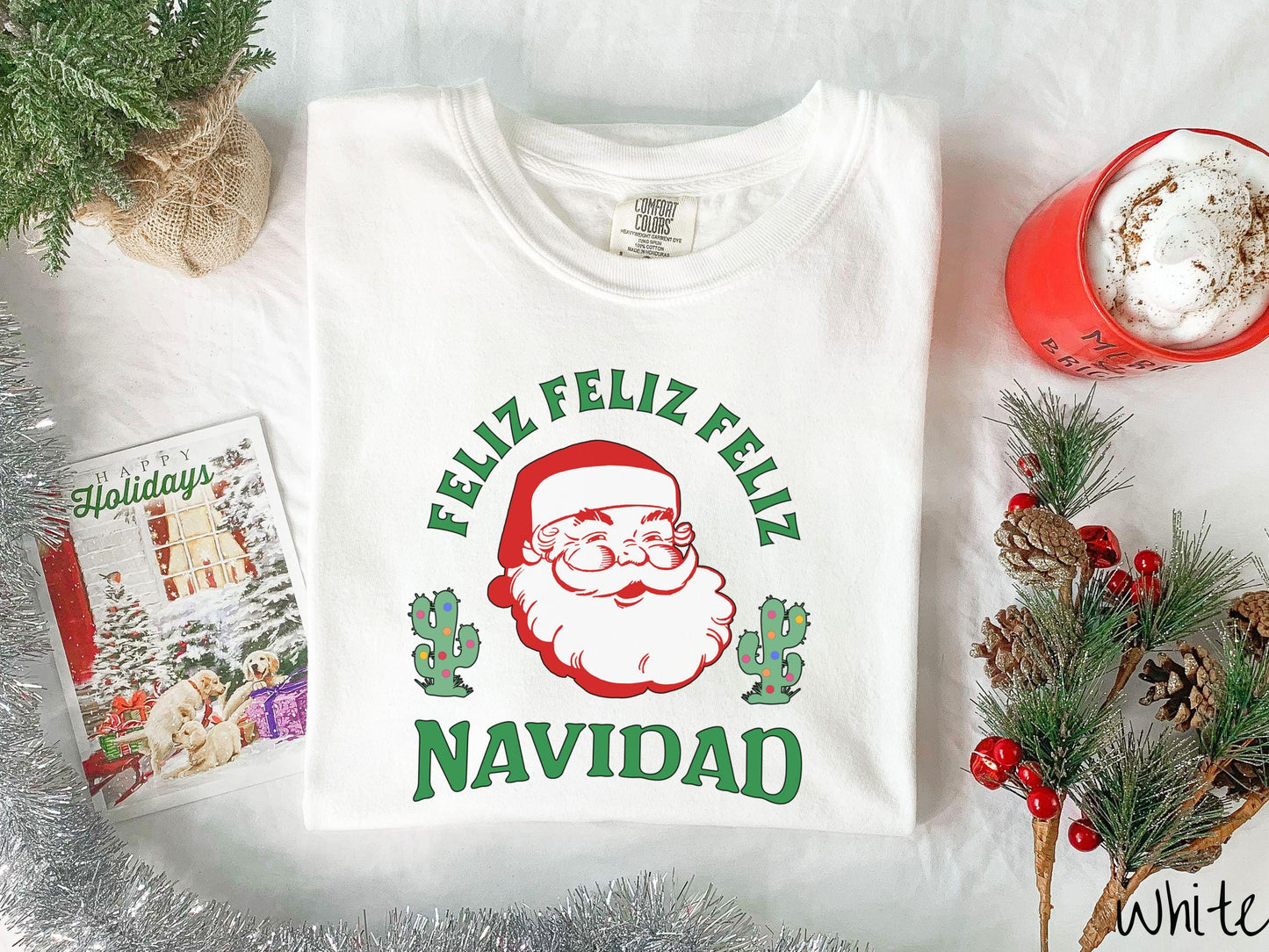 This festive white colored Comfort Colors t-shirt featuring a smiling santa clause between two green cacti covered in colorful Christmas lights and the phrase, 'Feliz feliz feliz Navidad' is perfect for spreading holiday cheer.