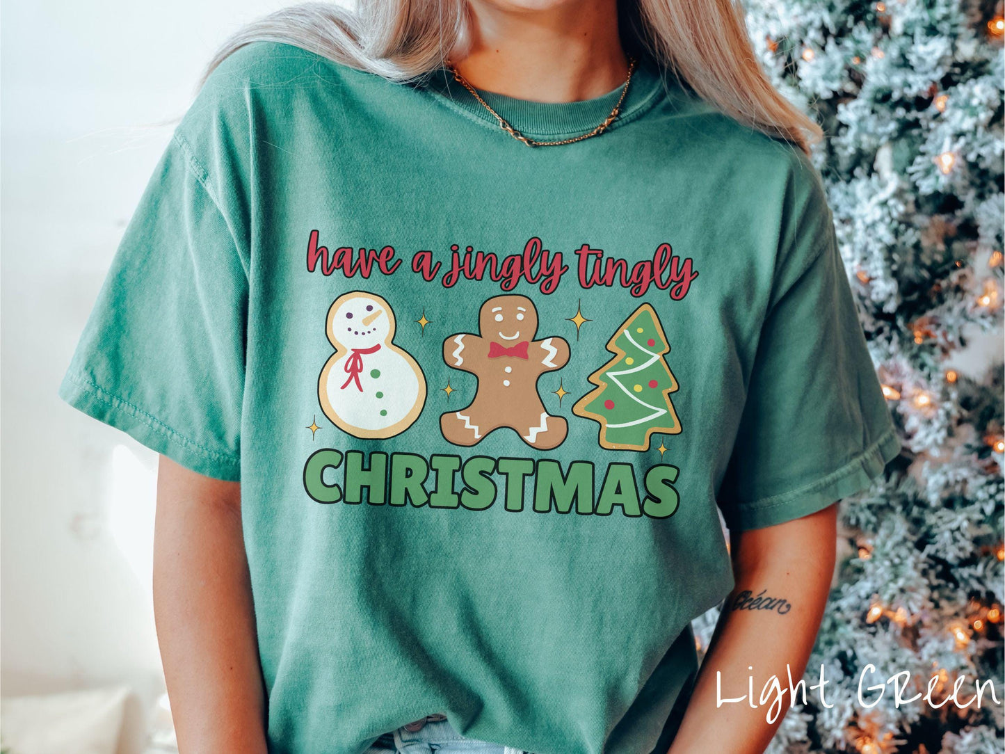 A vintage light green colored Christmas Comfort Colors t-shirt featuring a snowman iced cookie, a gingerbread man cookie, and a Christmas tree cookie. The text reads 'have a jingly tingly Christmas' in red and green font.
