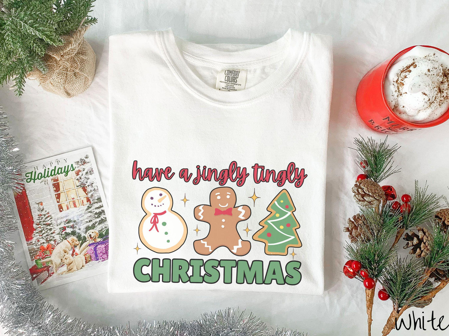 A vintage white colored Christmas Comfort Colors t-shirt featuring a snowman iced cookie, a gingerbread man cookie, and a Christmas tree cookie. The text reads 'have a jingly tingly Christmas' in red and green font.