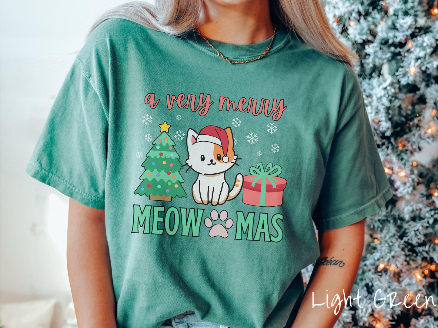 A vintage light green colored Comfort Colors T-shirt with the text a very merry Christmas in red and green font, featuring a grey cat wearing a red Santa hat sitting between a Christmas tree and a present with white snowflakes hanging in the air.