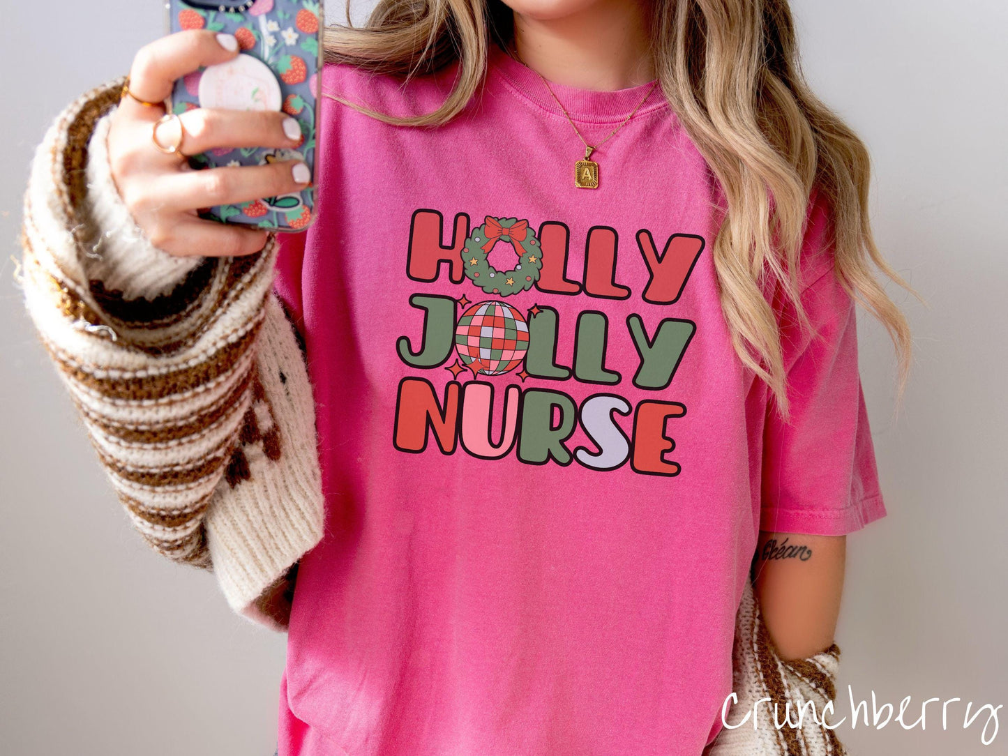 A woman wearing a cute, vintage crunchberry colored Comfort Colors T-shirt with text Holly Jolly Nurse in red, green , pink, and blue font. The letter O in Holly is a Christmas wreath and the letter O in Jolly is a red, green, and blue disco ball.