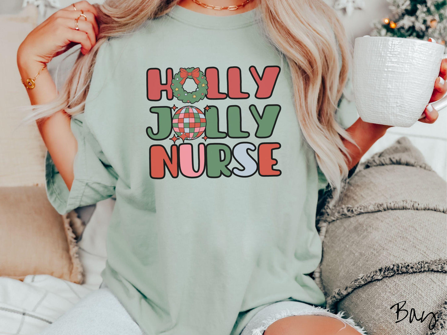 A woman wearing a cute, vintage bay colored Comfort Colors T-shirt with text Holly Jolly Nurse in red, green , pink, and cadet blue font. The letter O in Holly is a Christmas wreath and the letter O in Jolly is a red, green, and blue disco ball.