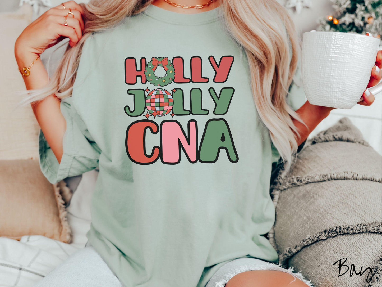 A woman wearing a cute, vintage bay colored Comfort Colors T-shirt with the text Holly Jolly CNA in red, green, pink, and blue font. The letter O in Holly is a Christmas wreath and the letter O in Jolly is a multi-colored disco ball.